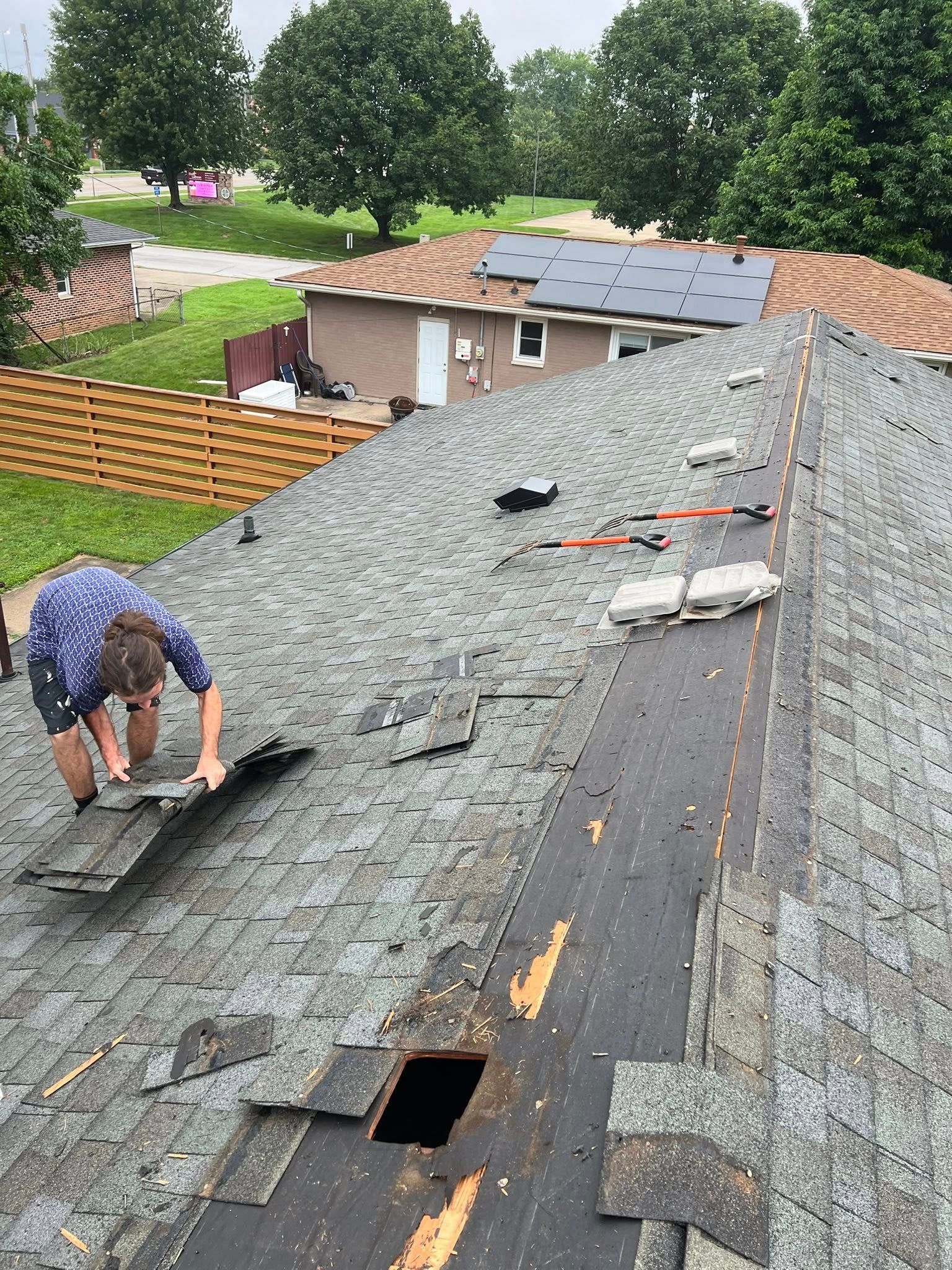 A person in a patterned shirt works on a residential roof, removing damaged shingles near an open vent.