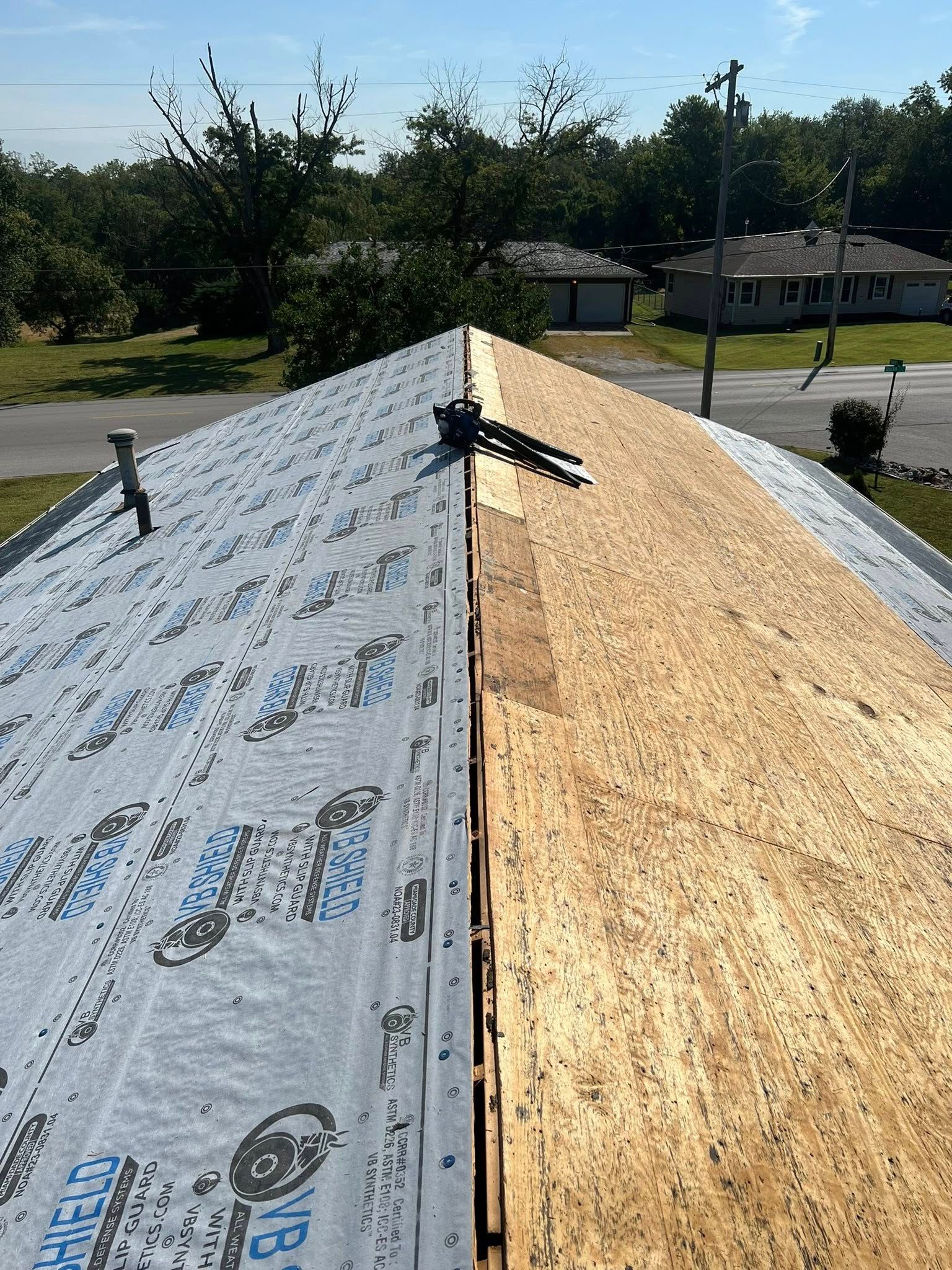 Roof under repair with gray underlayment on the left and exposed plywood on the right in bright sunlight.