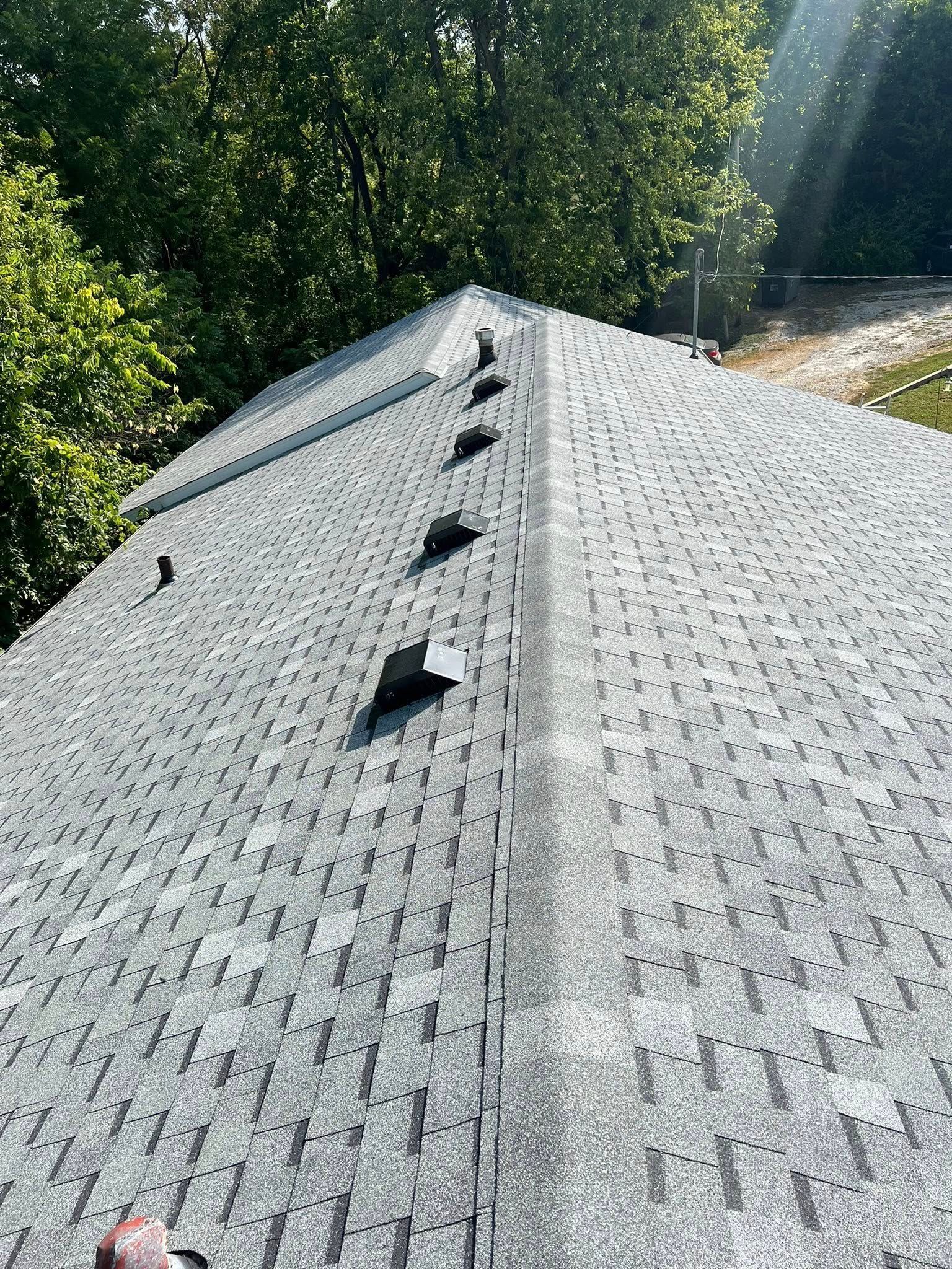 Sunlit gray shingle roof with vents, viewed from the ridge against leafy trees