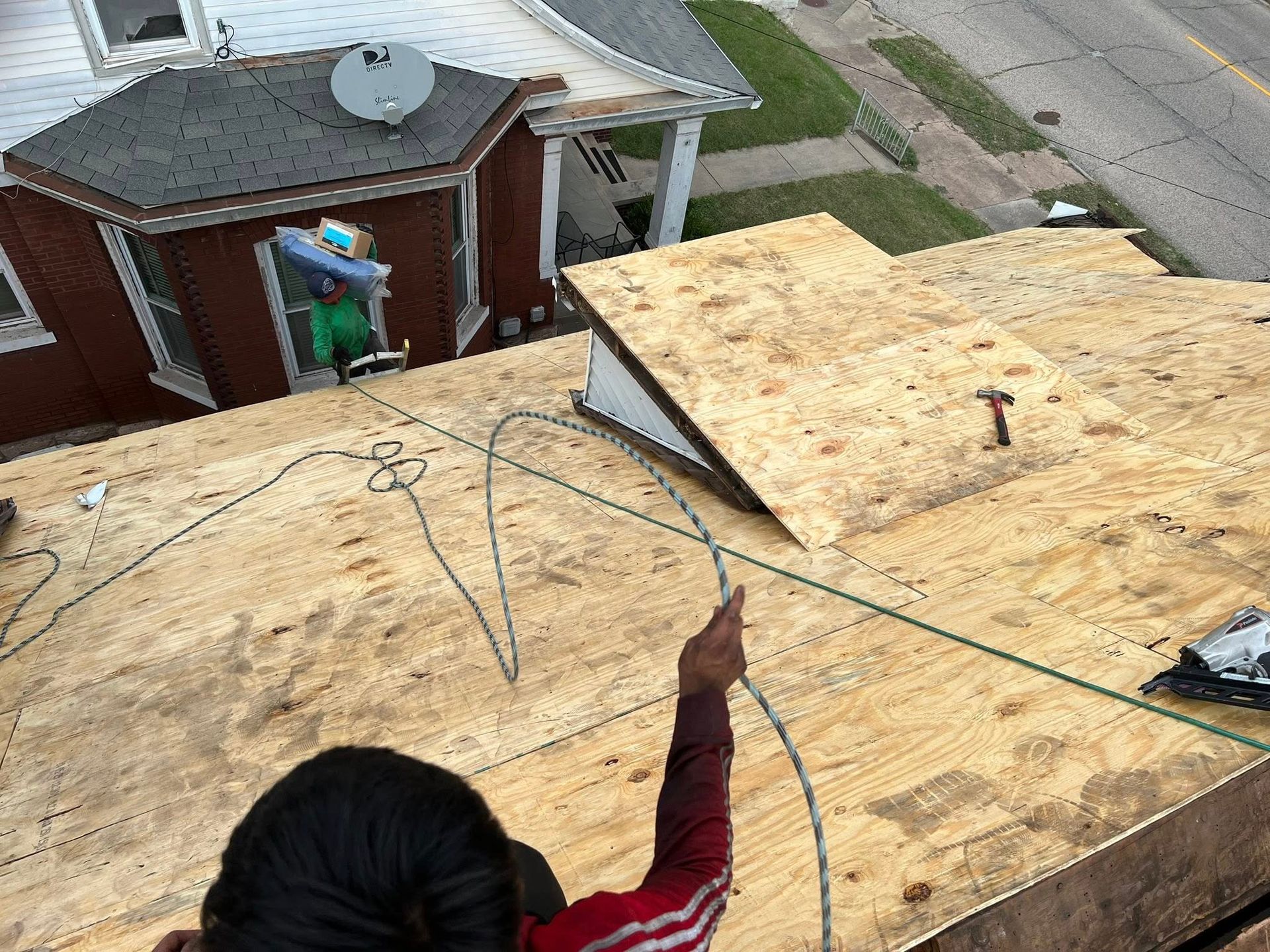 A person on a sloped roof under construction points toward a raised plywood section while holding a rope tether.