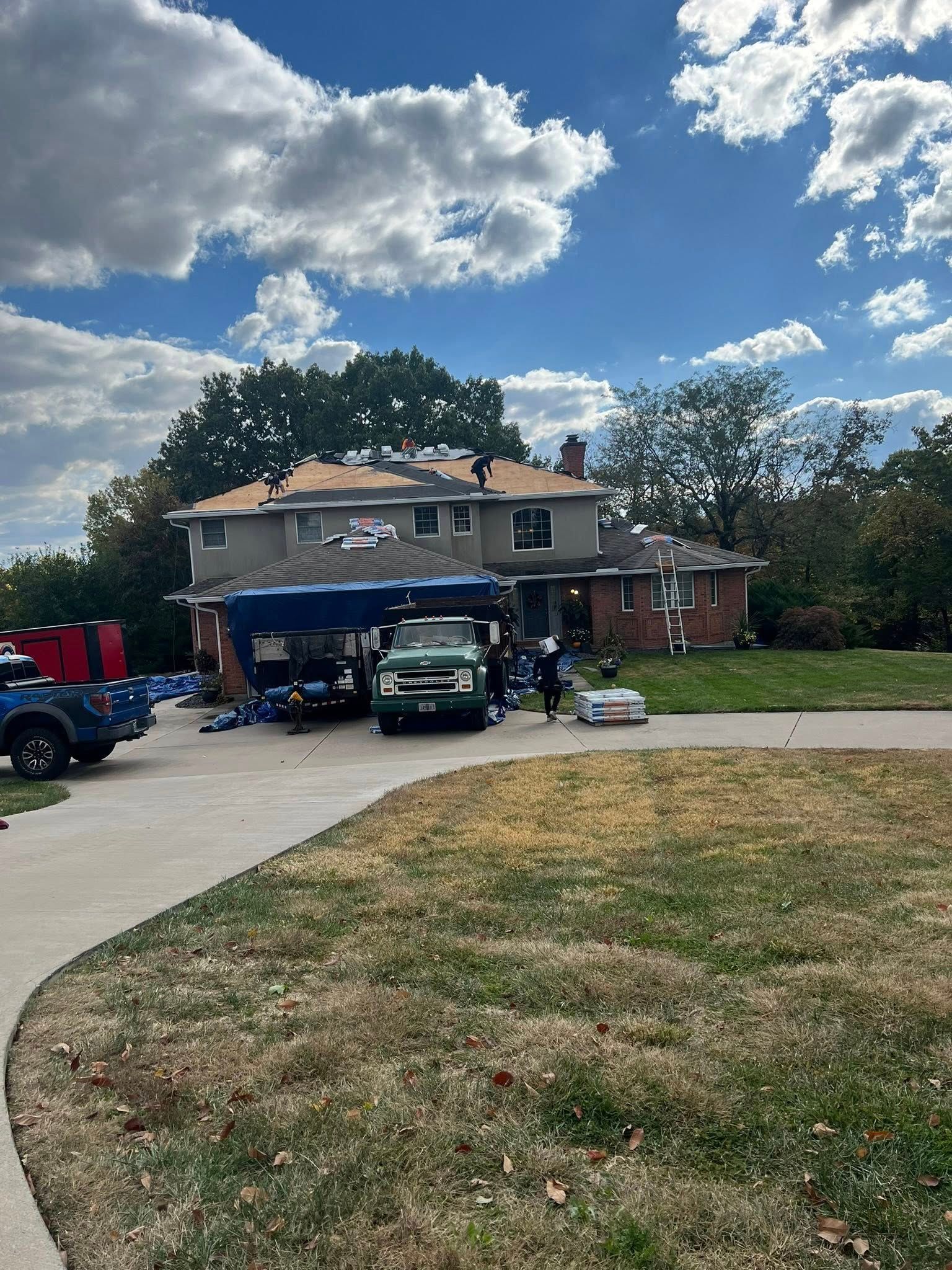 Suburban house with cars in the driveway, people gathered under the carport, and a cloudy blue sky above.