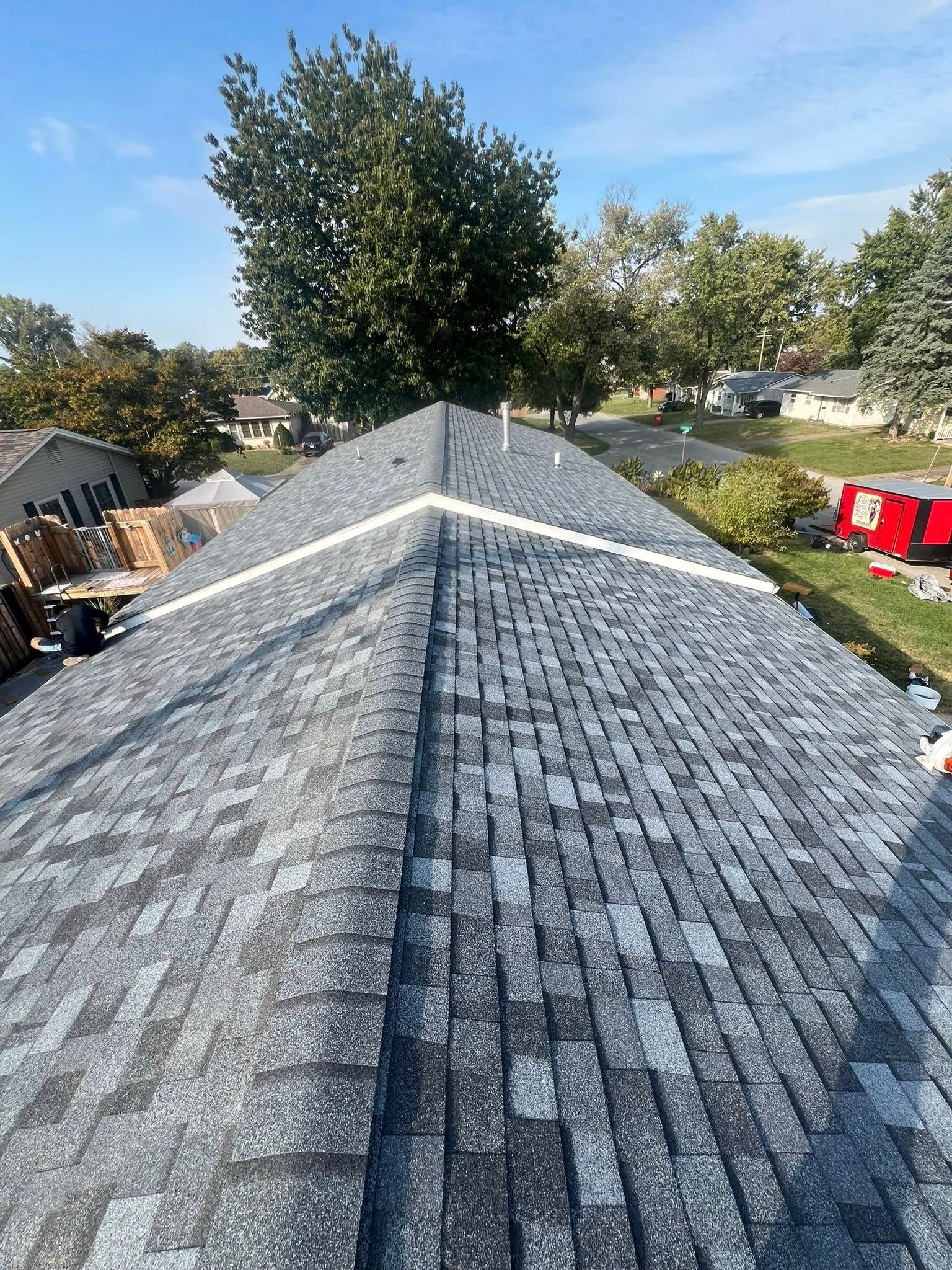 Gray shingle roof ridge under a blue sky, viewed from above with trees and yard equipment nearby