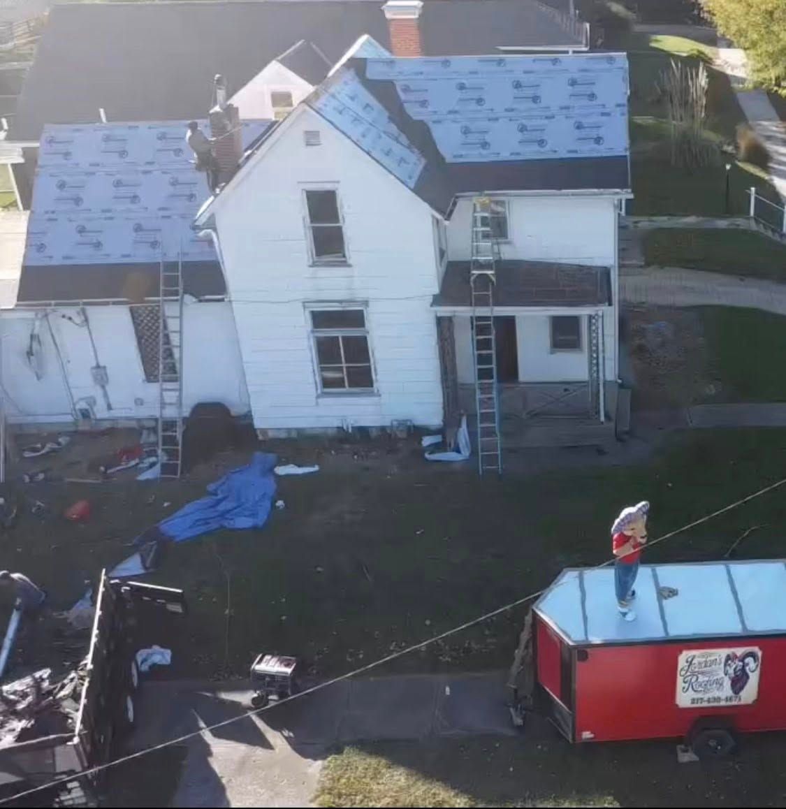 House under roof repair with ladders, tarps, and a red trailer in the yard