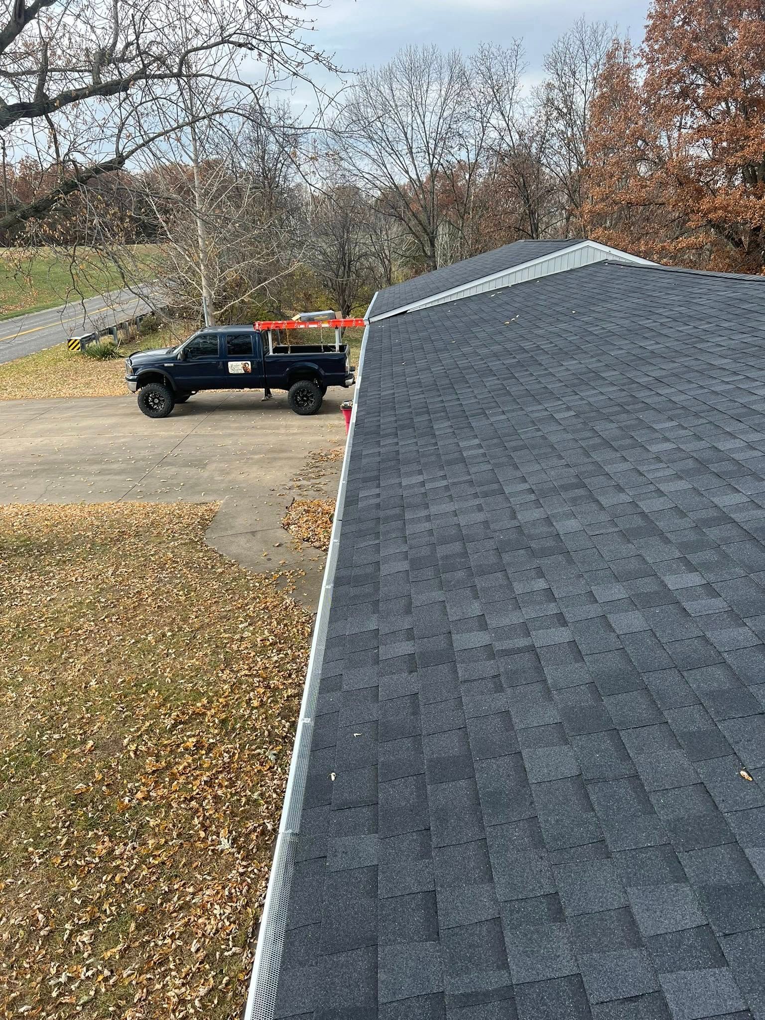 Roof shingles in foreground, with a black utility truck parked on a driveway beside a leaf-covered yard.