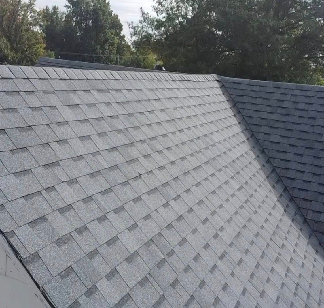 Gray shingled roof sloping beside a darker roof section with trees in the background