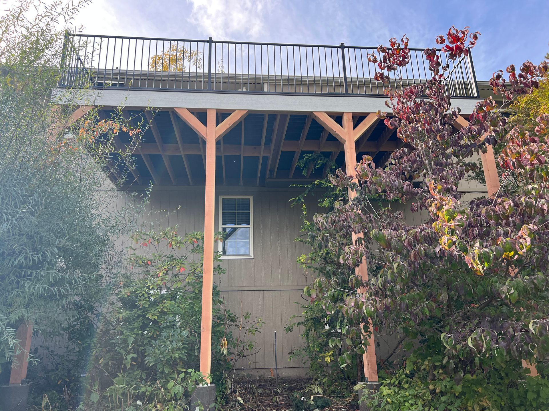 Wooden deck supported by posts, over a tan-colored building with a window and door, surrounded by greenery.