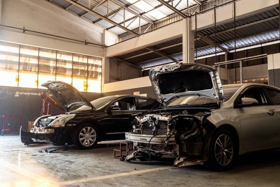Two cars with open hoods in a dim auto repair garage, one front bumper removed.