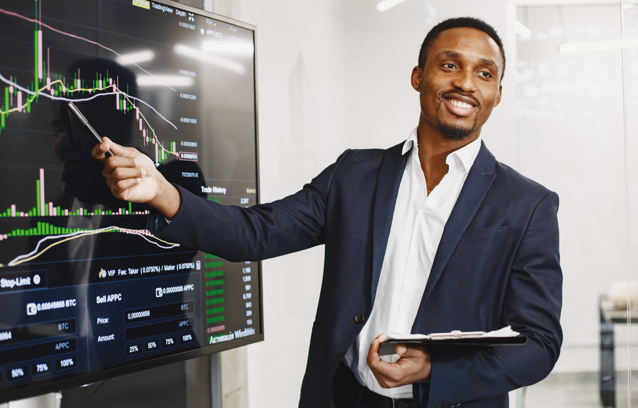 Man in suit points at a financial chart on a large screen, smiling.