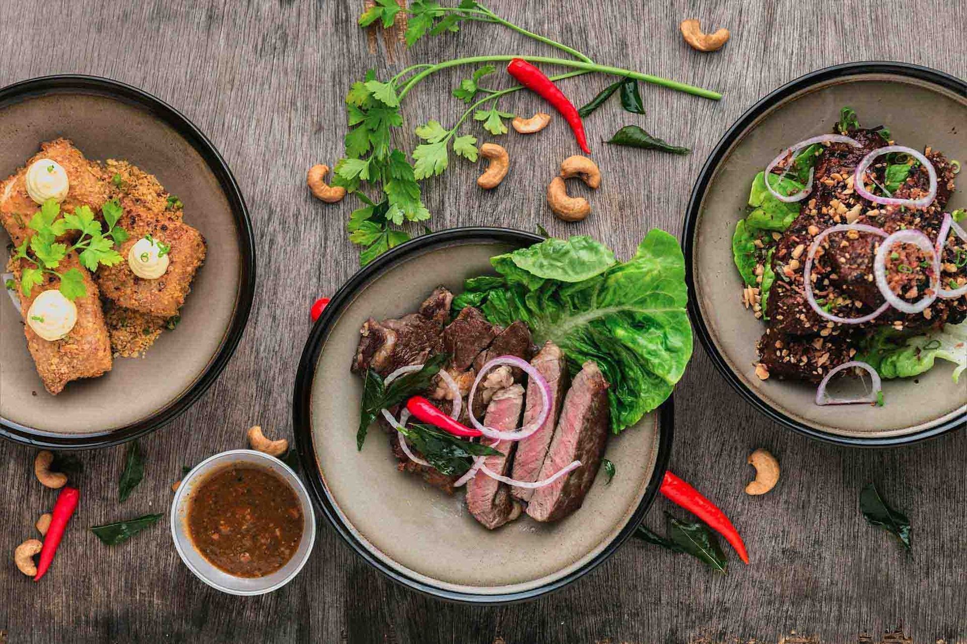 Three rustic bowls of sliced meat, herbs, and dipping sauce on a wooden table with red chilies