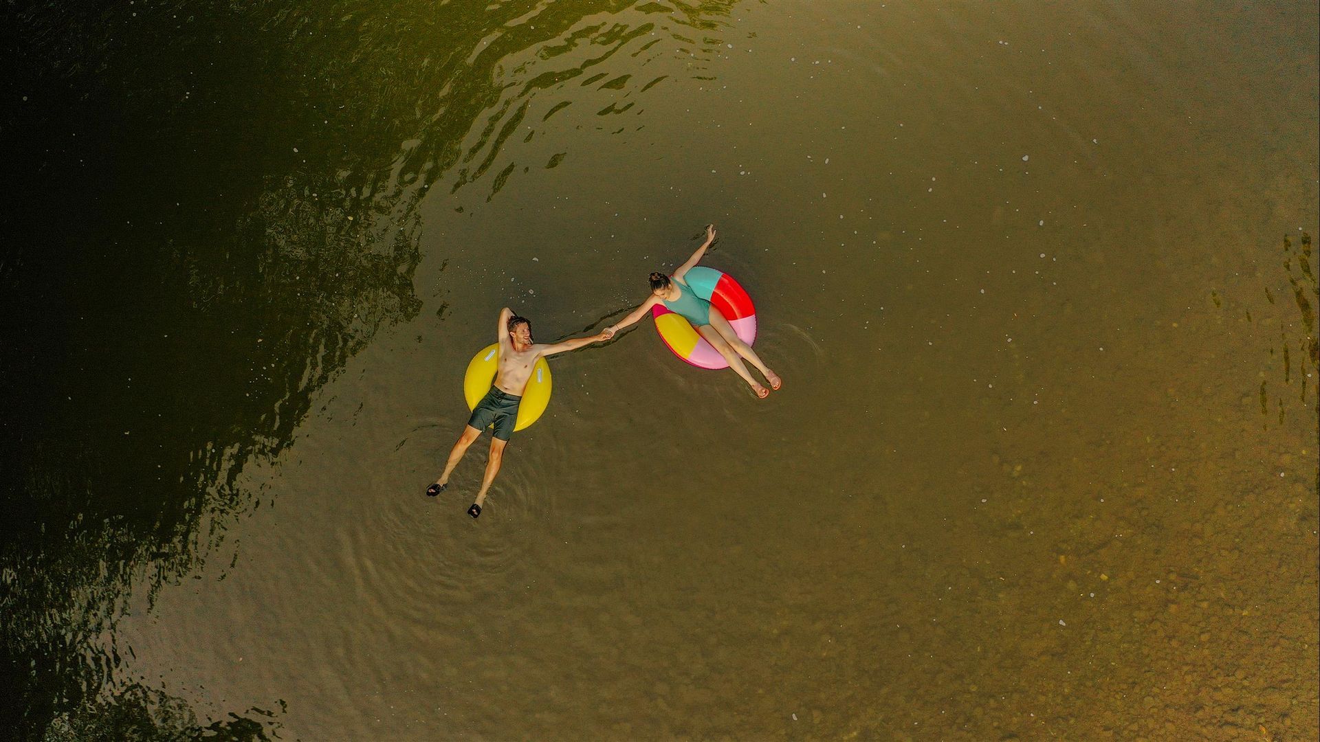 Two people in colorful life vests float on a calm river in a small boat, seen from above.