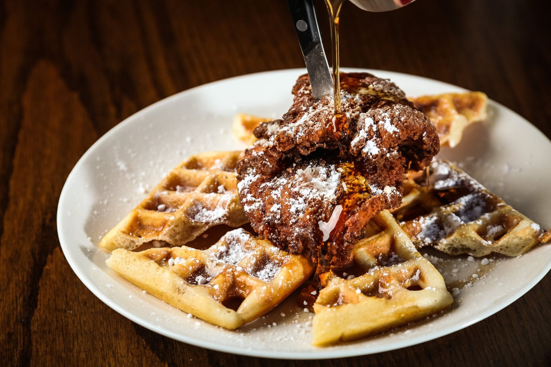 Waffles topped with fried chicken and powdered sugar on a white plate being cut with a fork and knife