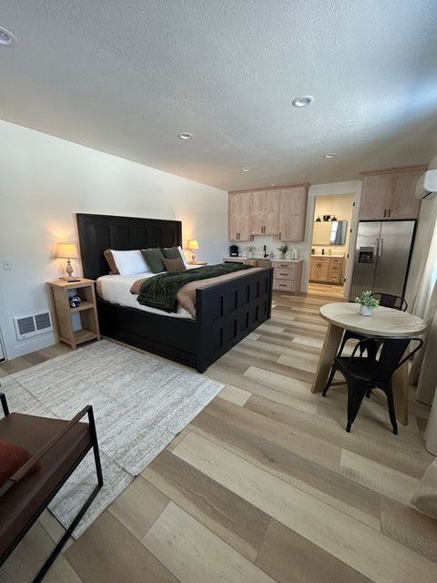 Modern bedroom with black bed, light wood flooring, small table, and kitchenette in the background