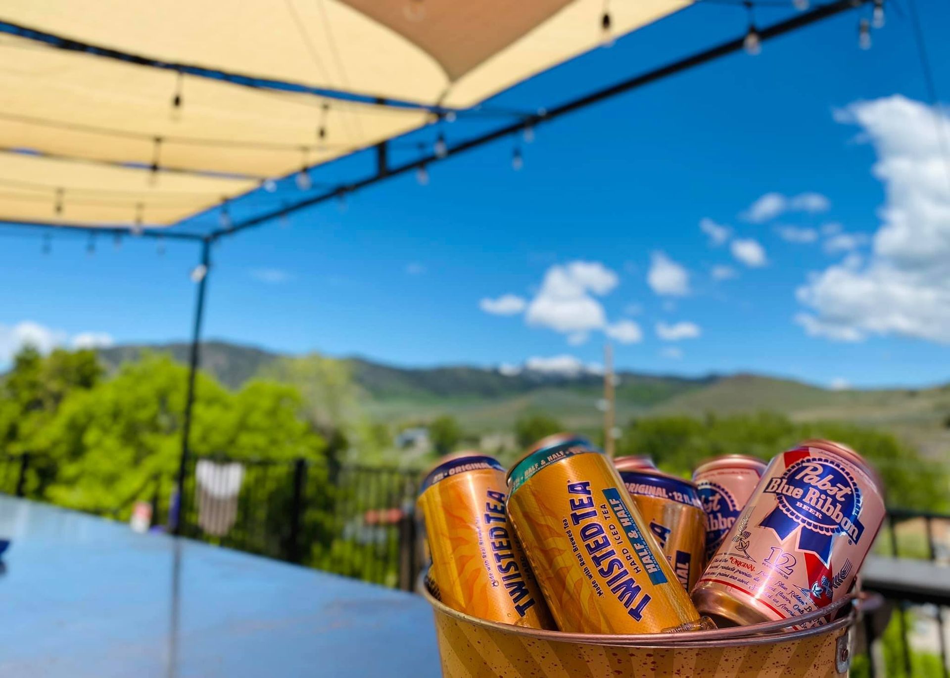 Basket of beer cans on an outdoor table under a canopy with a blue sky and hills in the background