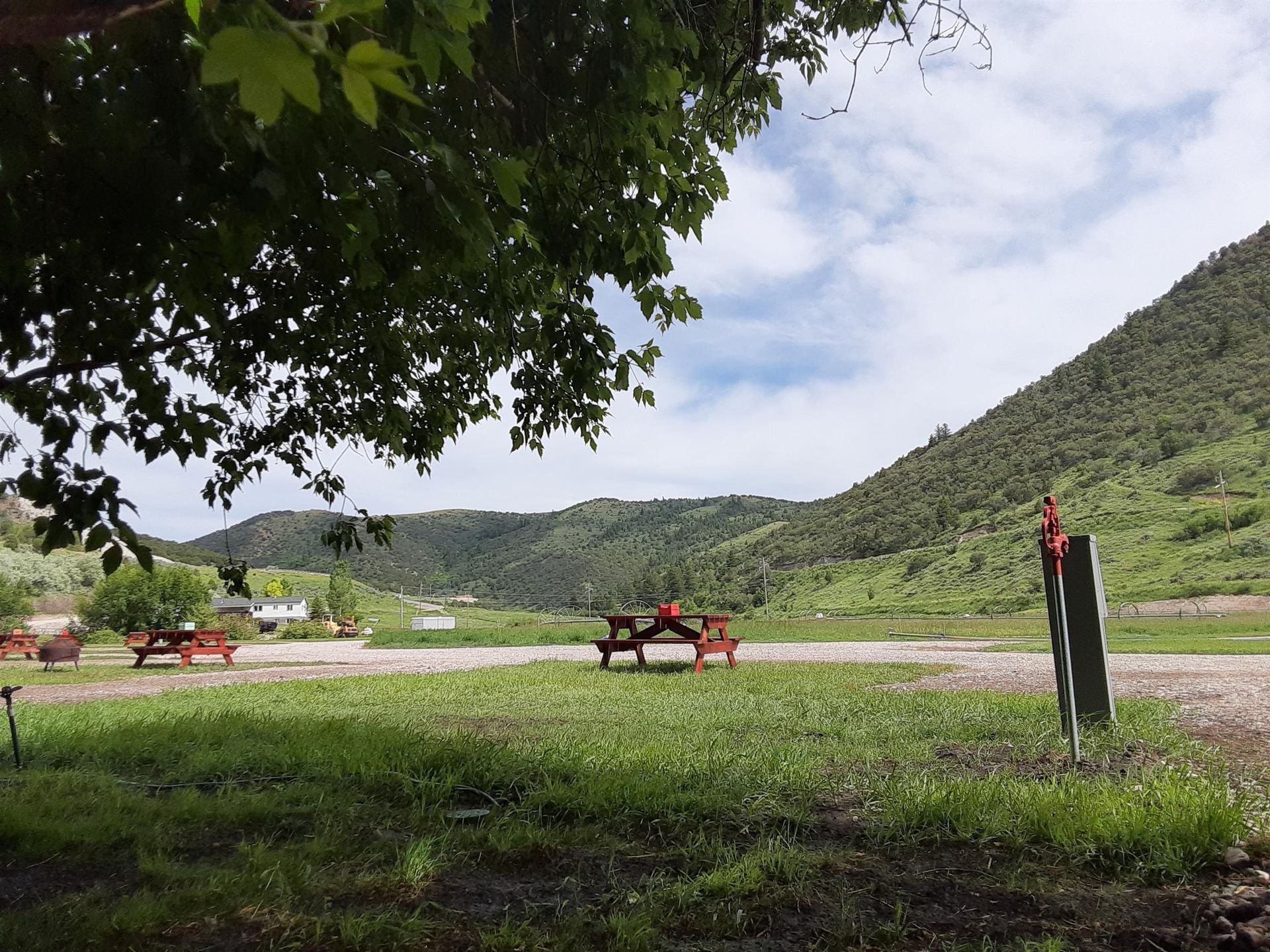Grassy field with picnic tables, a road, and green hills under a partly cloudy sky