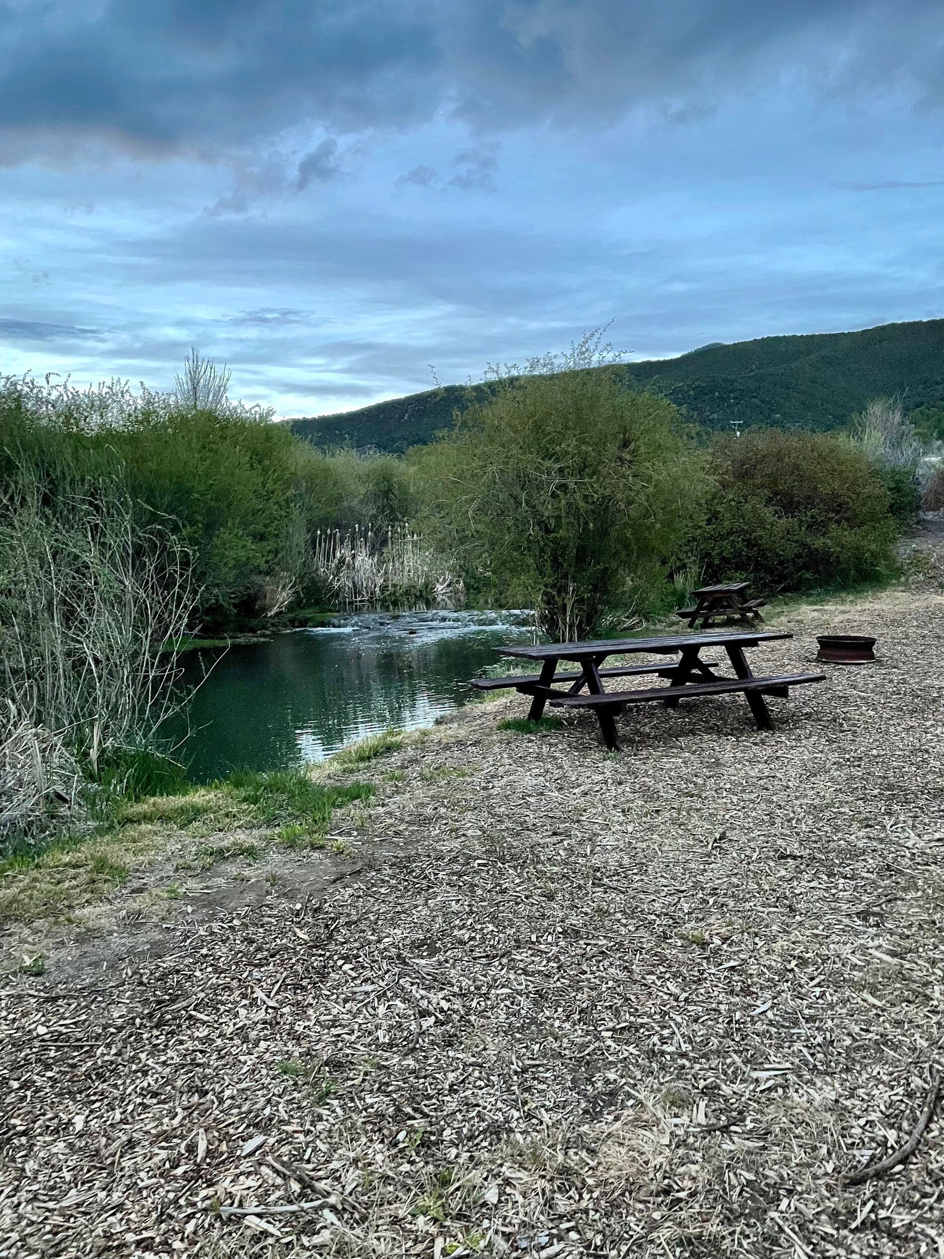 Gravel picnic area beside a calm river, with picnic tables and green hills under a cloudy sky.
