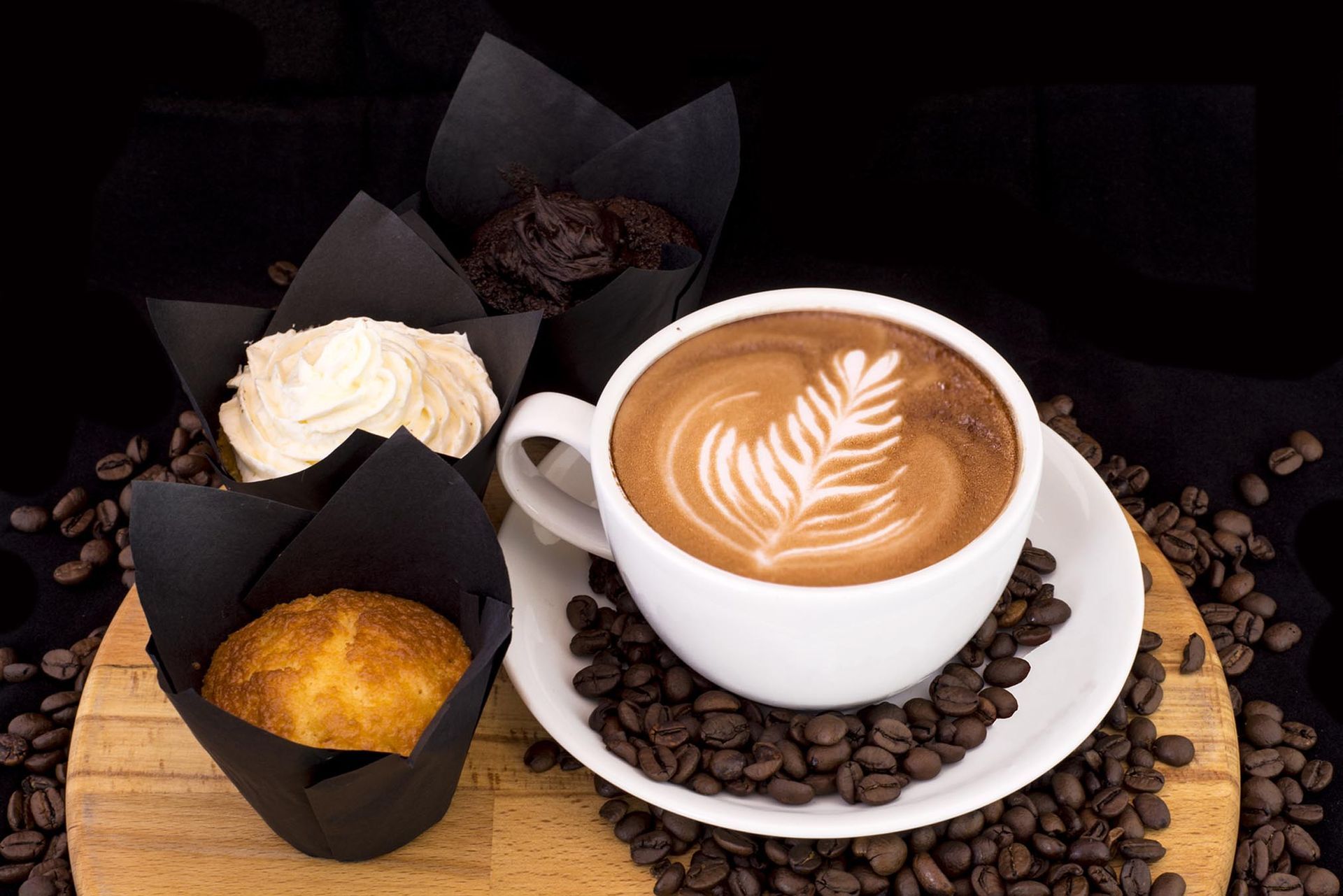 White latte in a cup with coffee beans and black paper-wrapped pastries on a wooden board