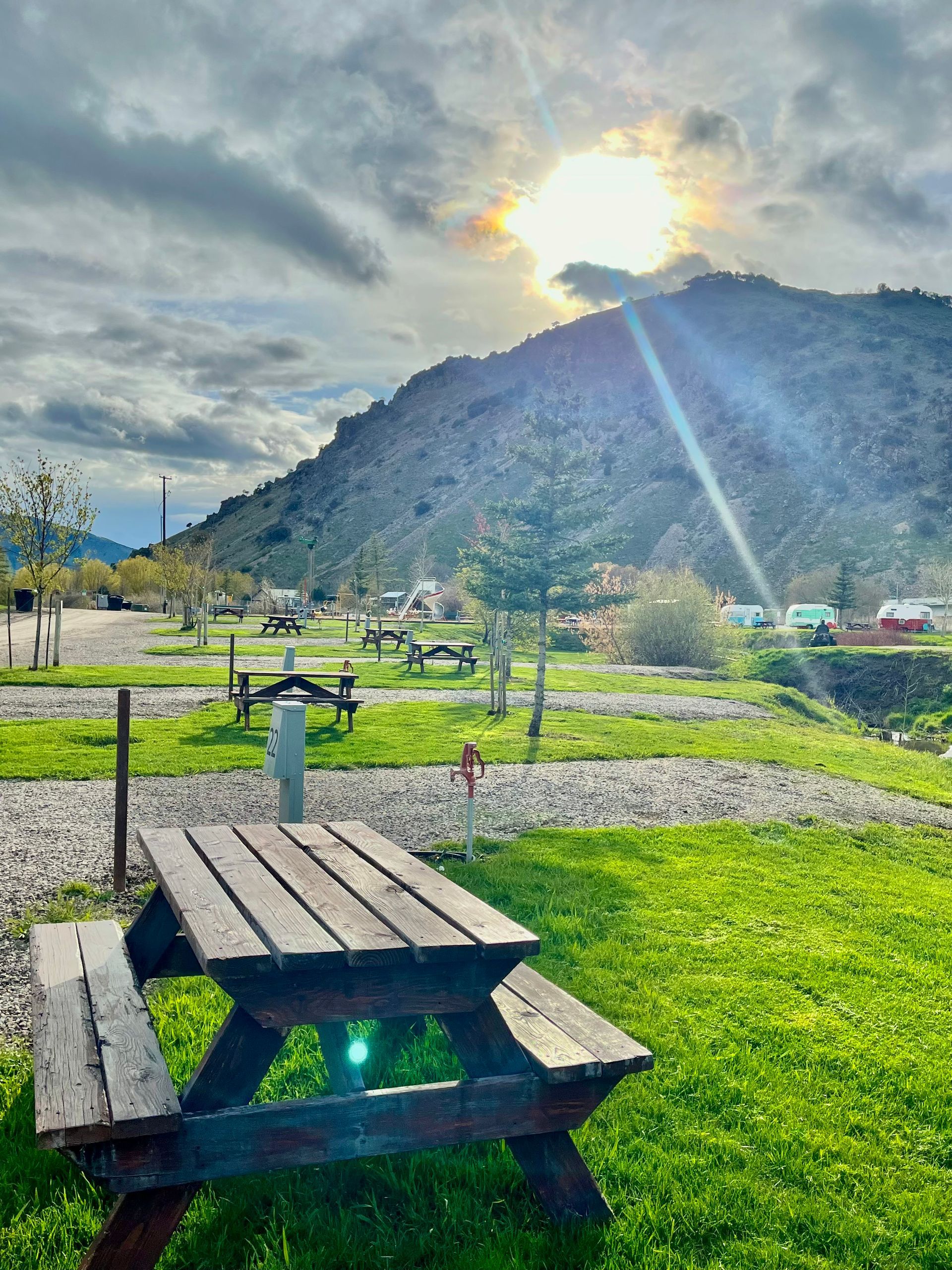 Wooden picnic table on green grass with a mountain and bright sun behind clouds in the background.