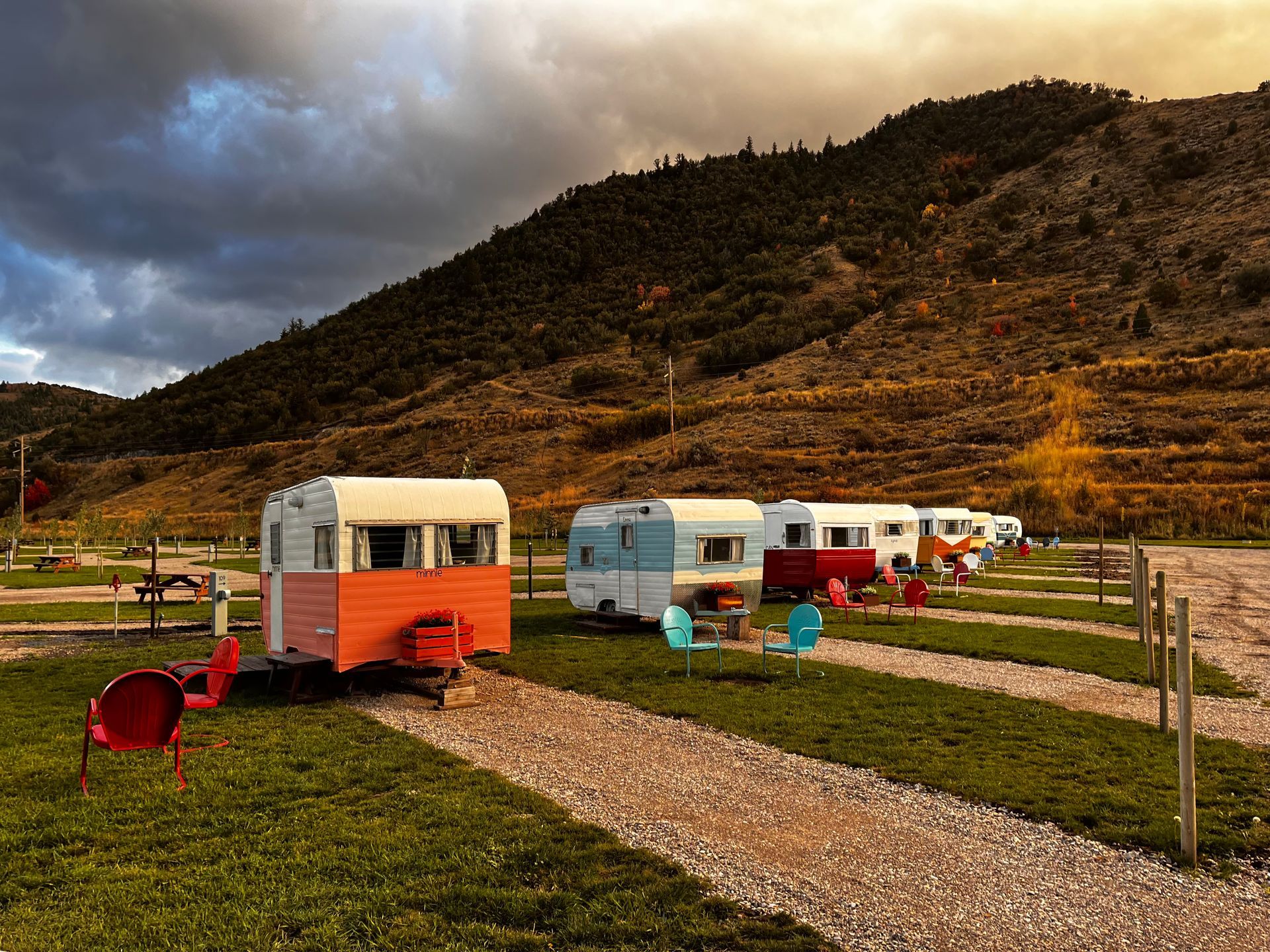 Small colorful campers beside gravel paths in a grassy field under a cloudy mountain sky