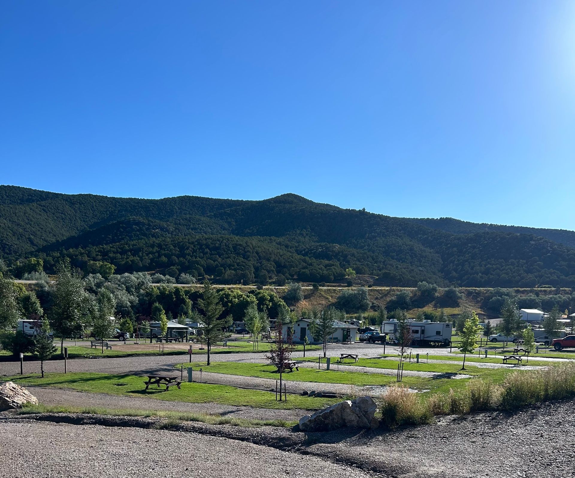 Sunlit mountain landscape with a grassy field, scattered trees, and a small town under a clear blue sky