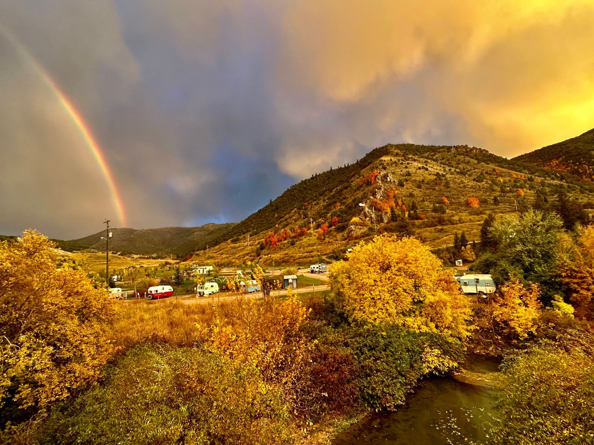 Autumn mountain landscape with golden trees, a rainbow, and a small village beside a river