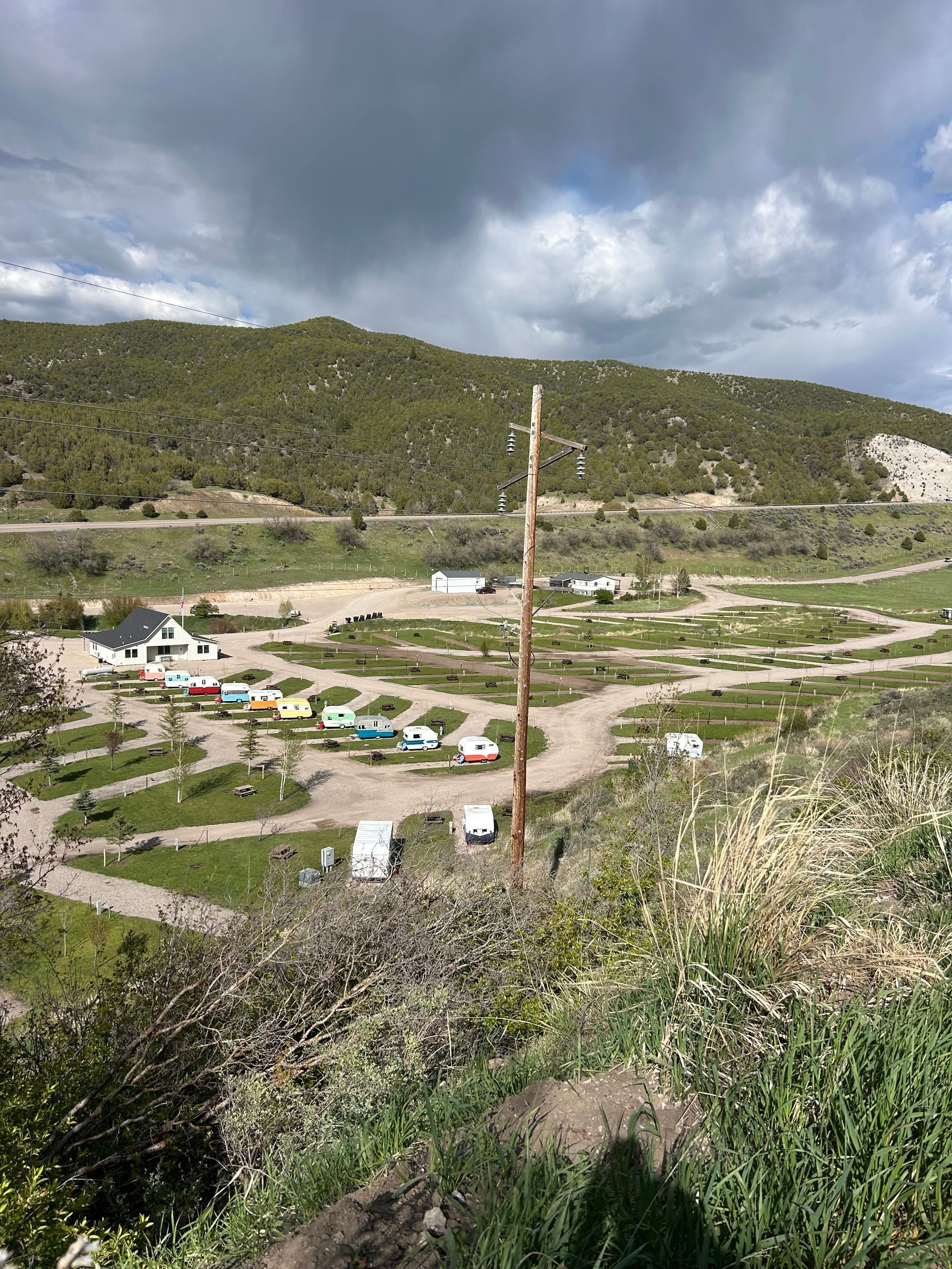Mountain valley with tents and small buildings under cloudy skies, viewed from a hillside with a tall pole in the foreground