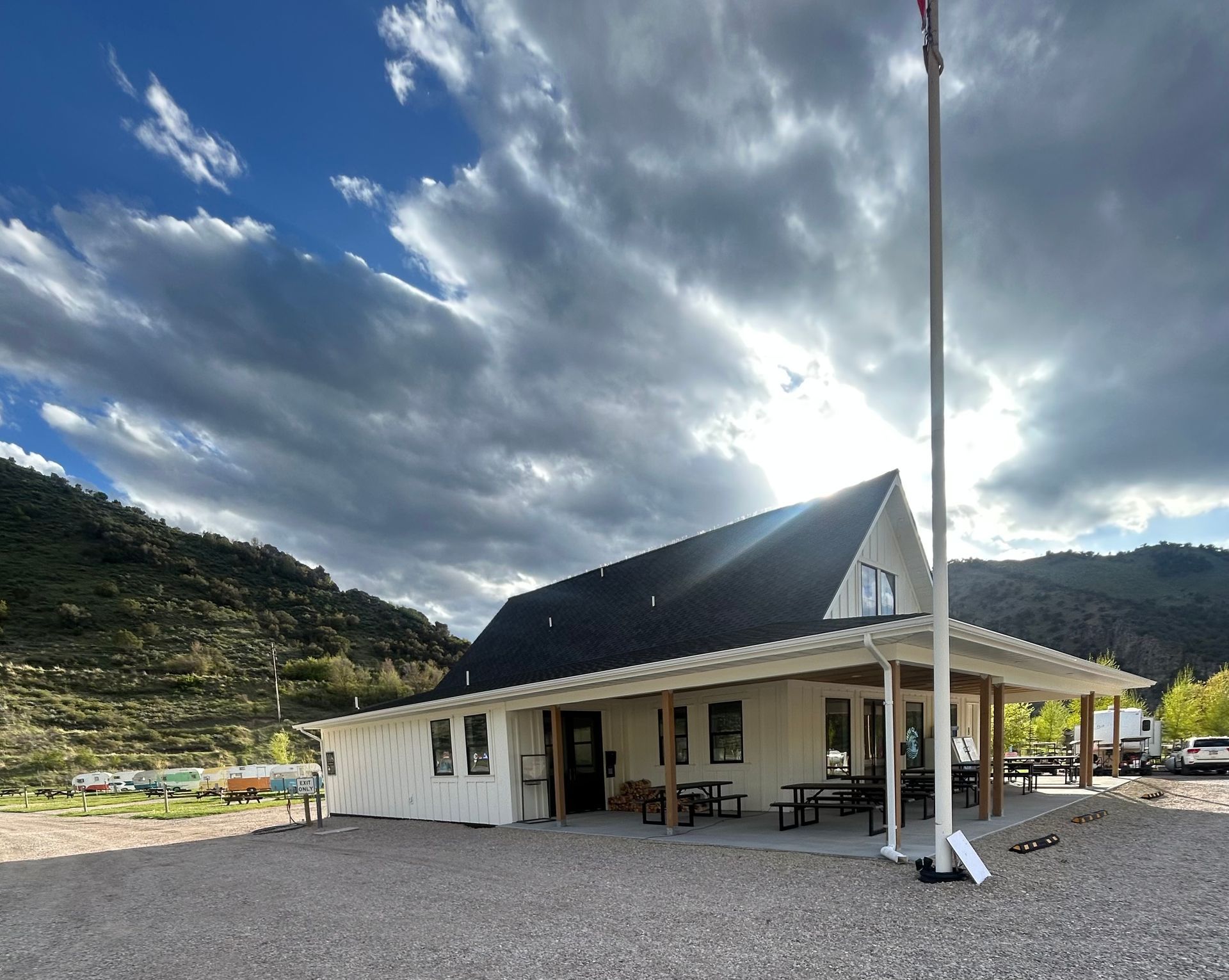 White building with dark roof on gravel, mountains behind, under a dramatic cloudy sky with bright sun rays
