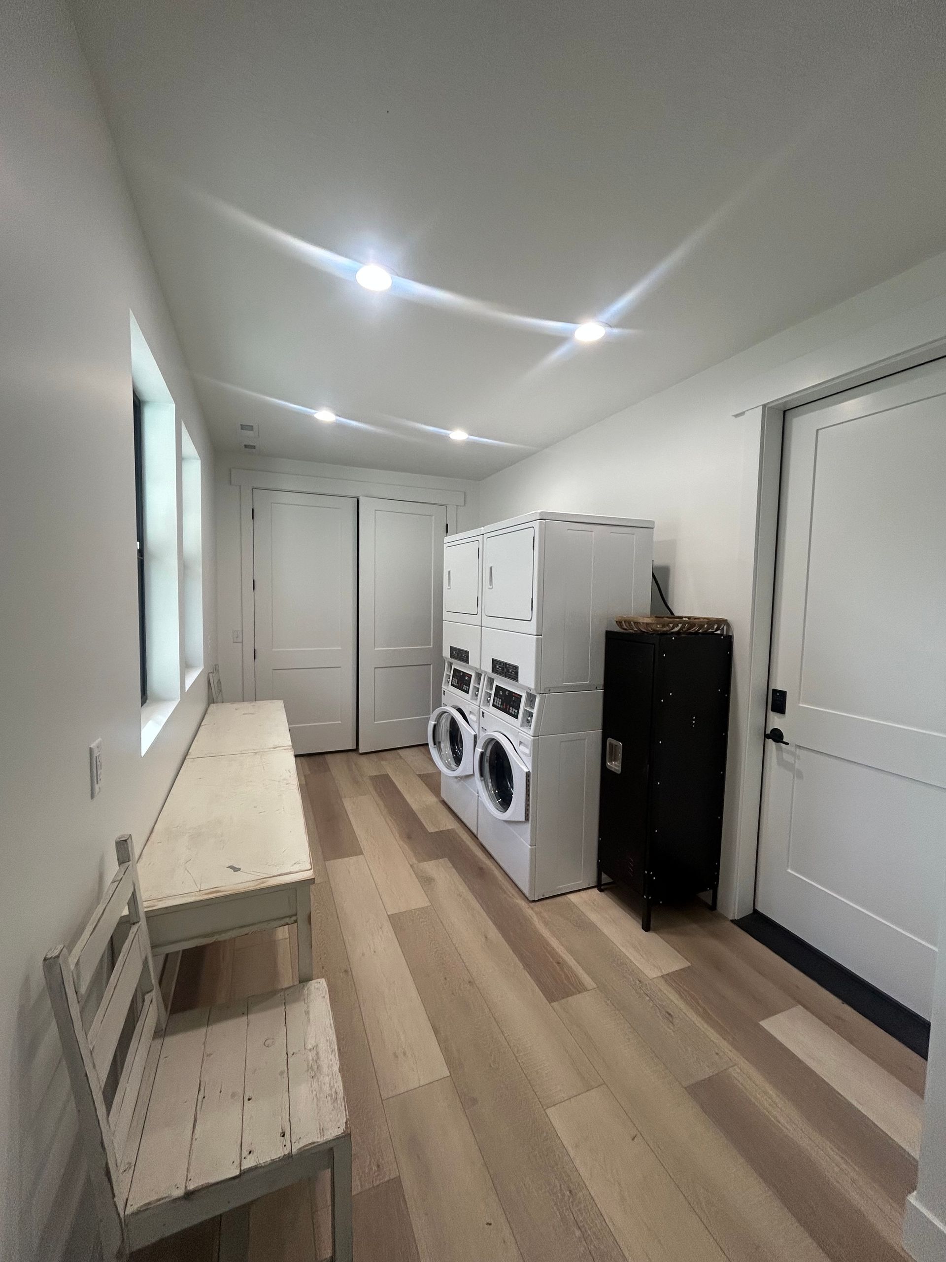 Narrow laundry room with stacked washer and dryer, white doors, and a bench along the left wall.