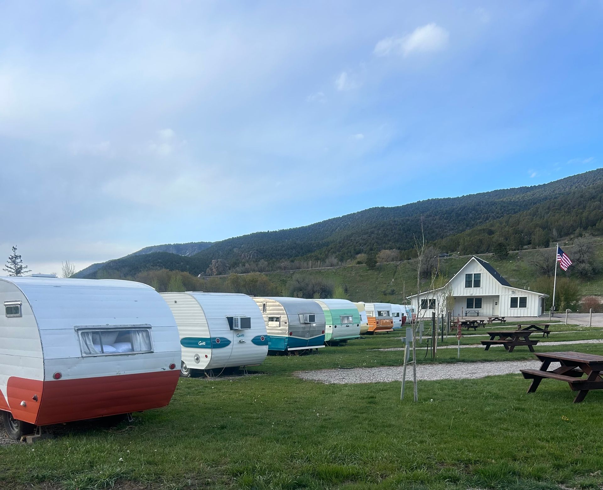 Camper trailers on a grassy field beside a house and picnic table, with mountains and blue sky behind.