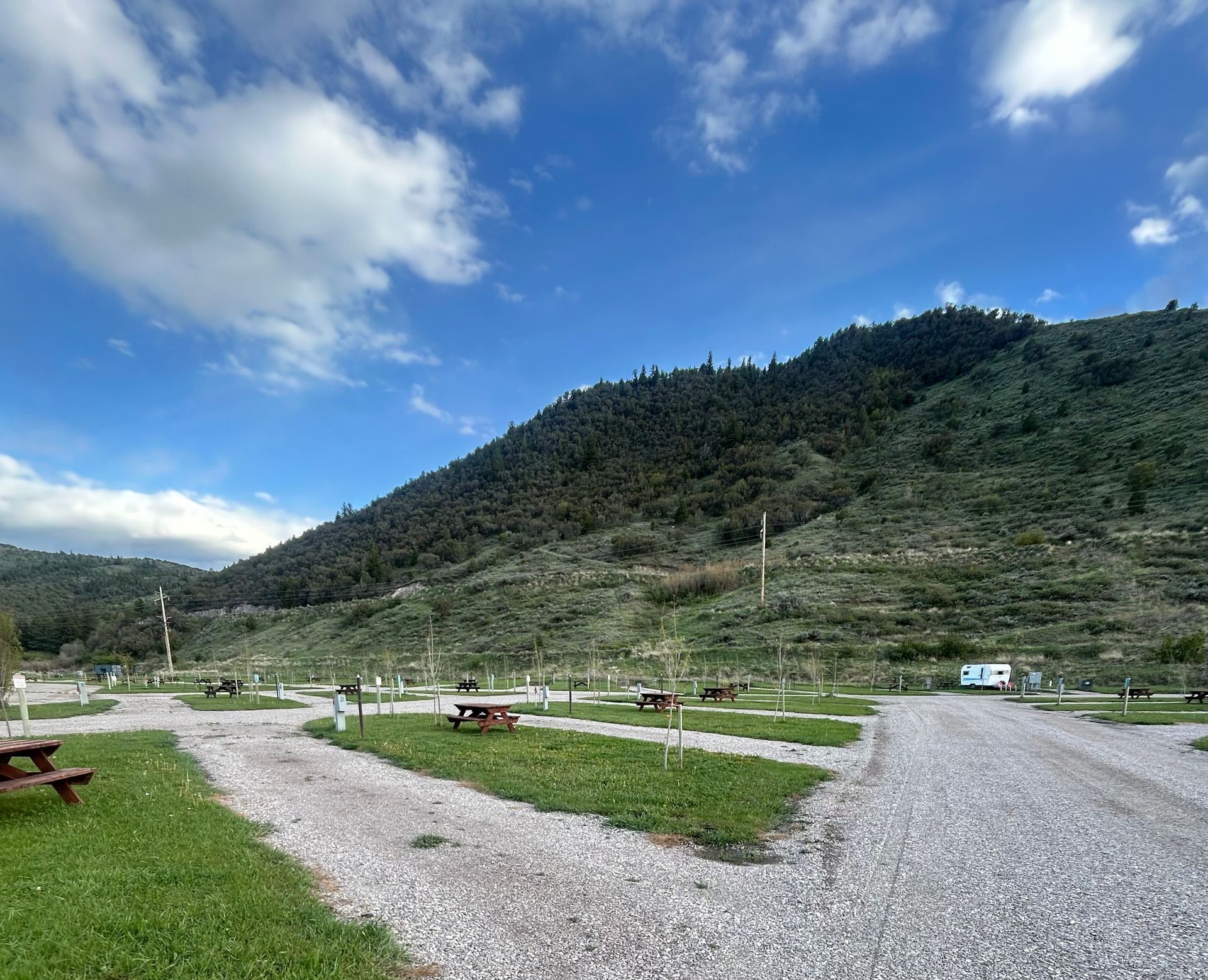 Gravel path and picnic tables in a green valley beneath a forested mountain under a blue, partly cloudy sky