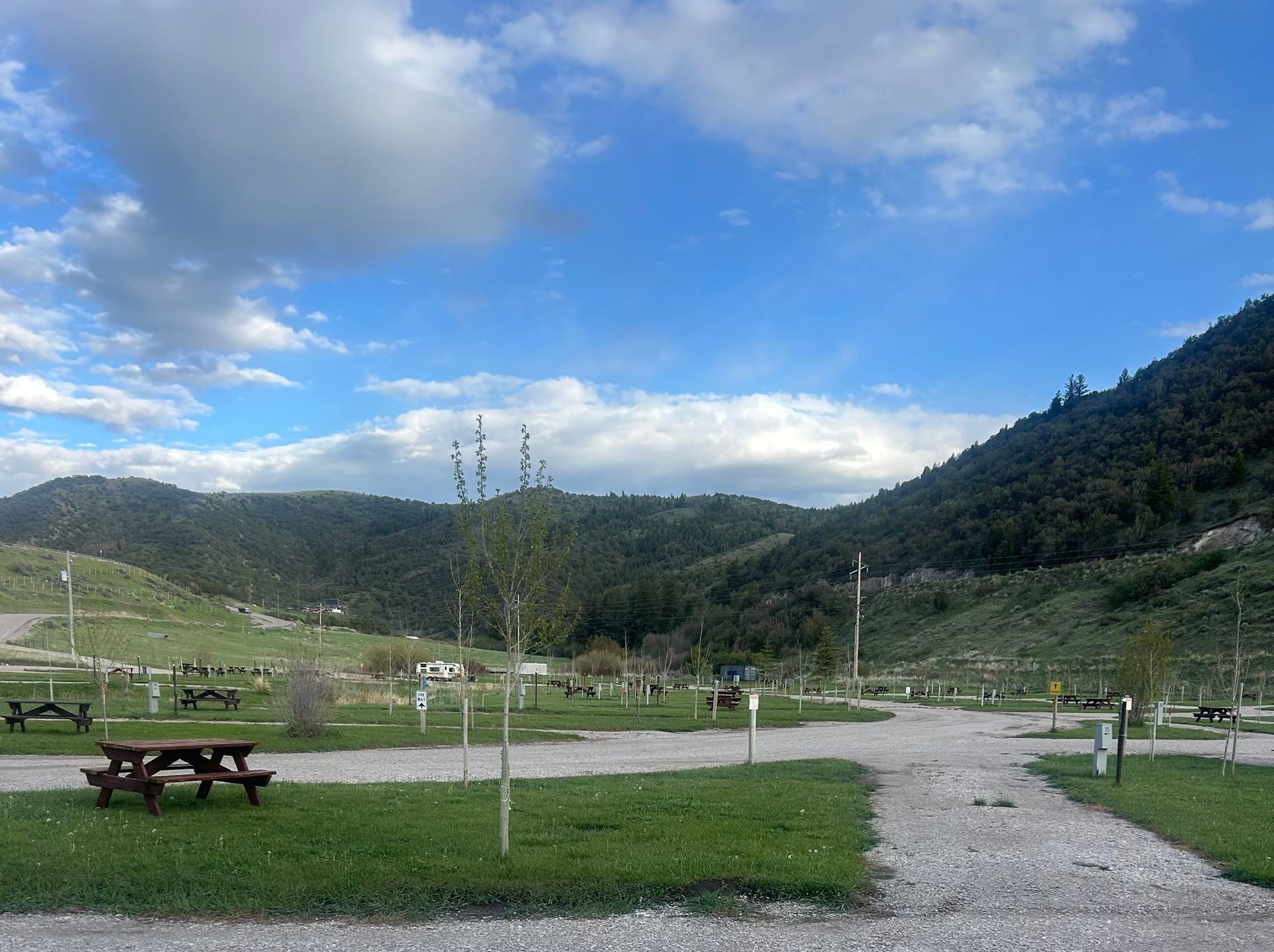 Grassy park with picnic tables, gravel path, and green hills under a cloudy blue sky