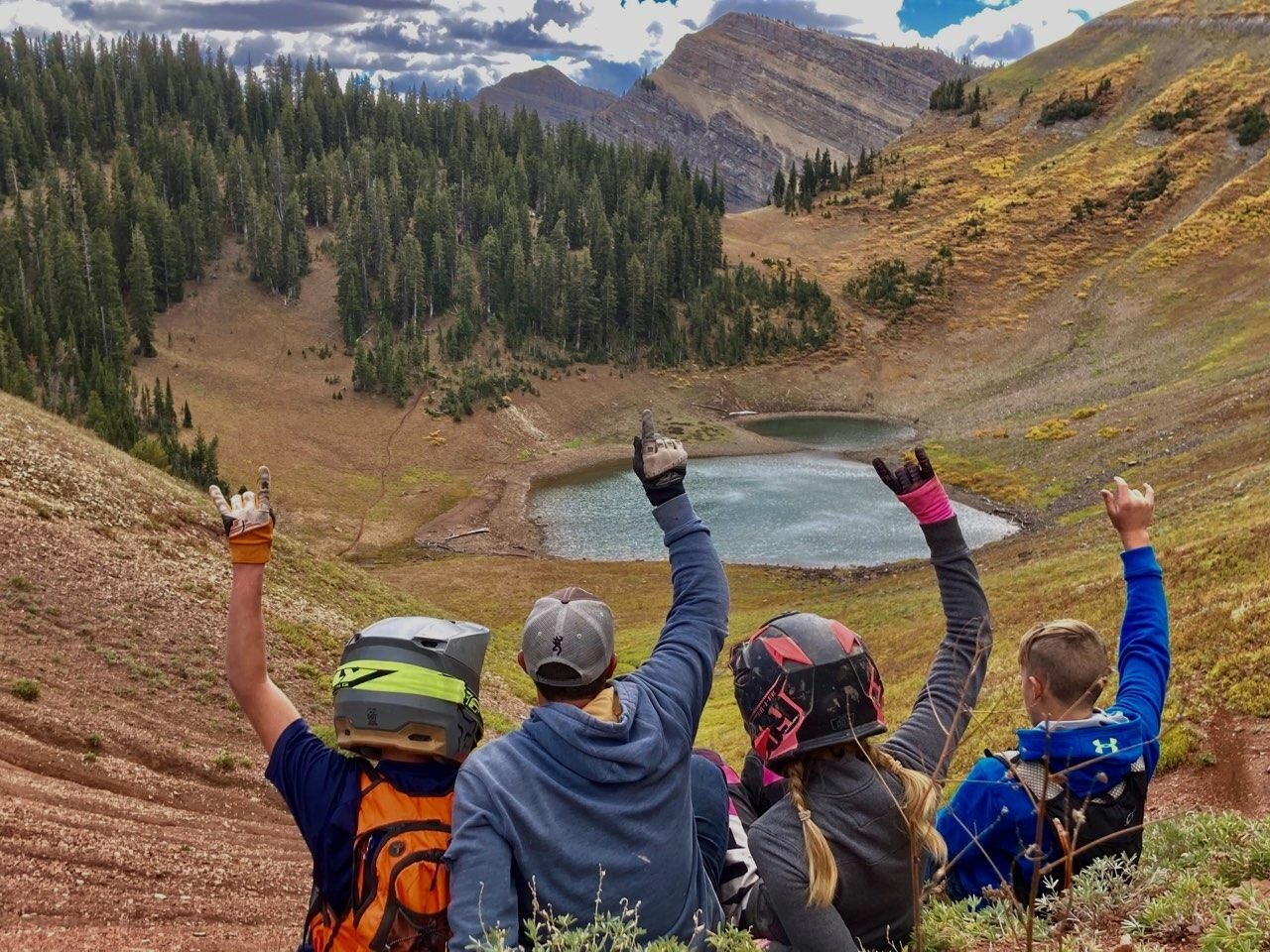 Four hikers with raised arms overlook a mountain lake and forested valley.