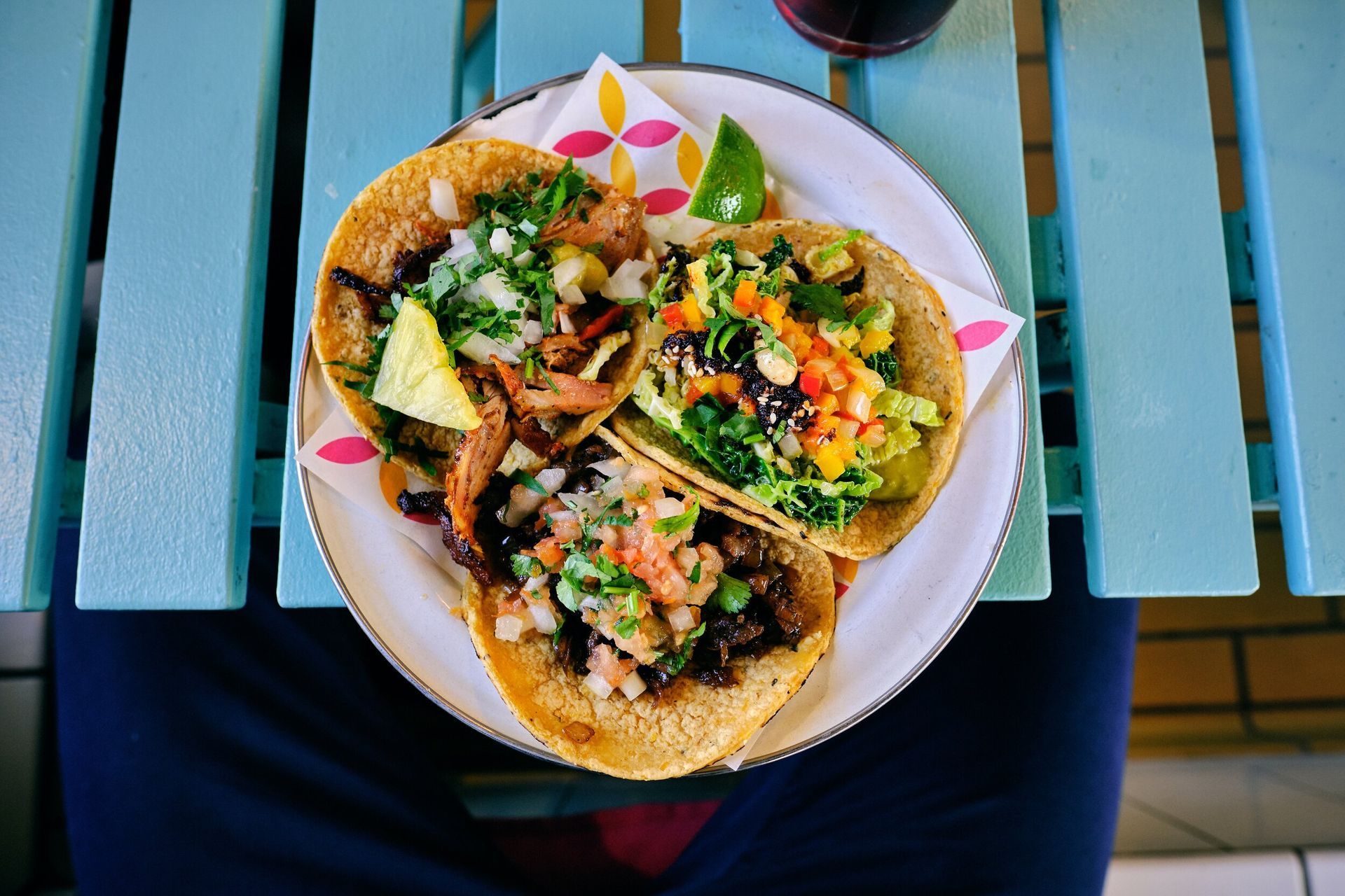 Three tacos on a white plate atop a person’s lap at a blue outdoor table