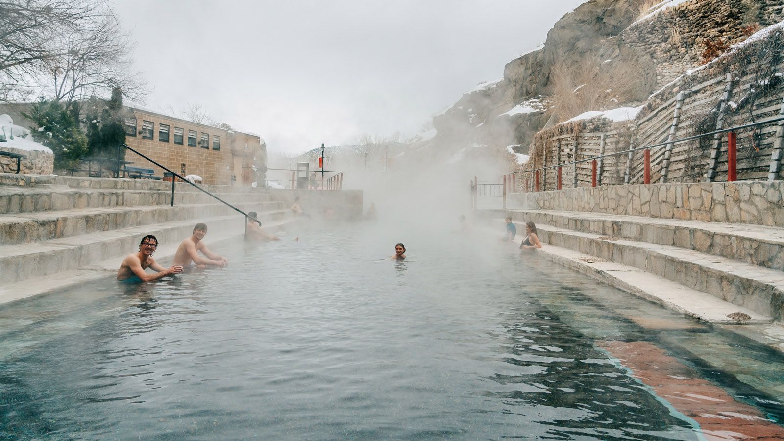 People soaking in a misty outdoor hot spring pool beside snow-covered stone terraces
