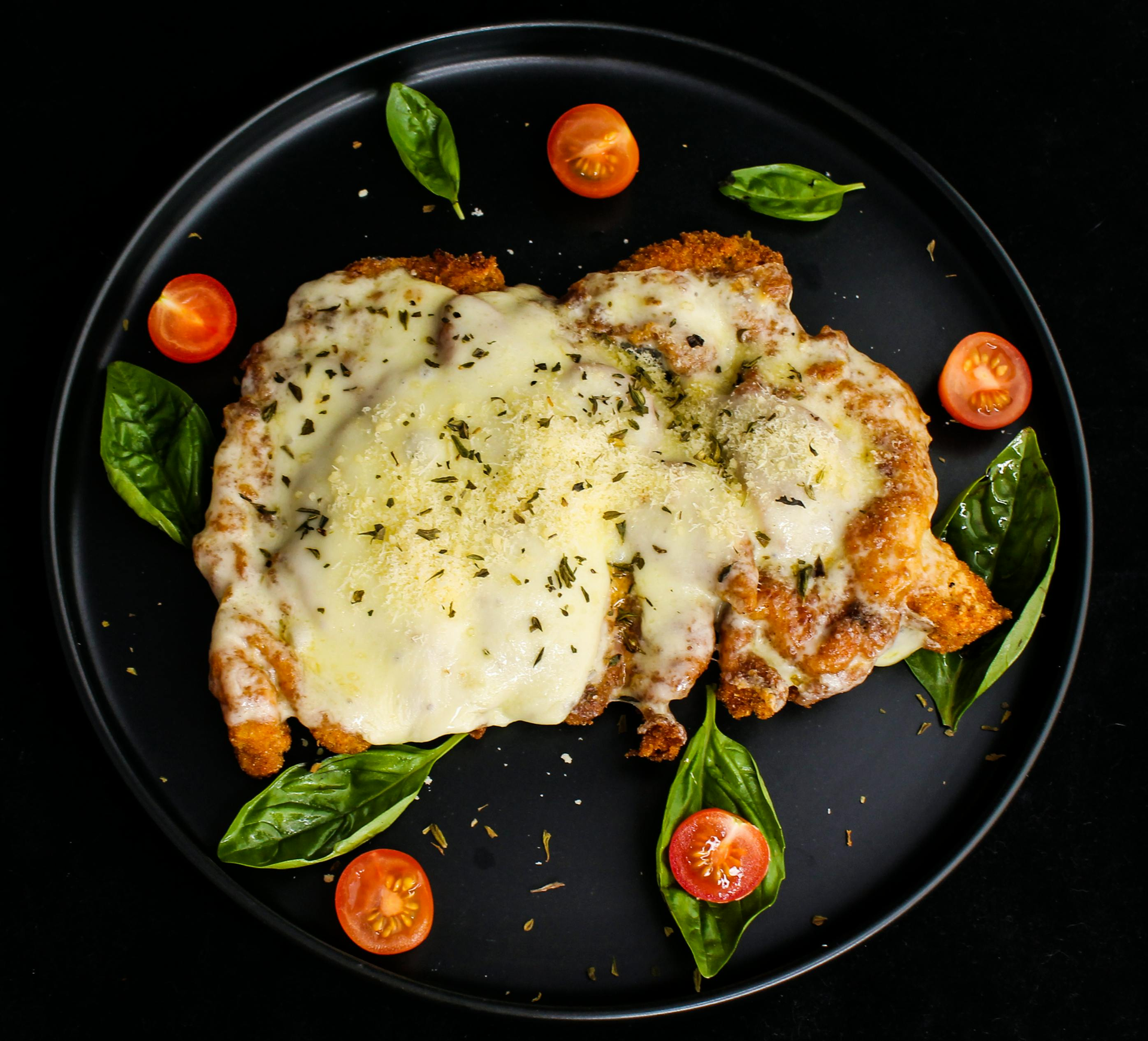 Melted cheese-covered baked chicken on a black plate, garnished with basil leaves and cherry tomatoes.