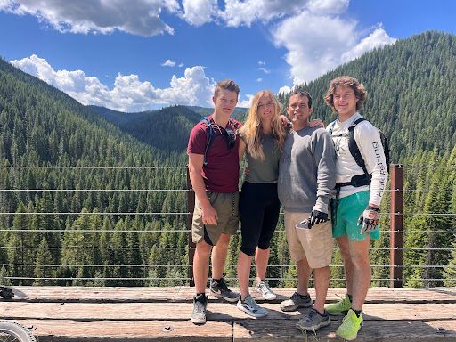 Three hikers standing on a wooden deck with forested mountains and a blue sky behind them.