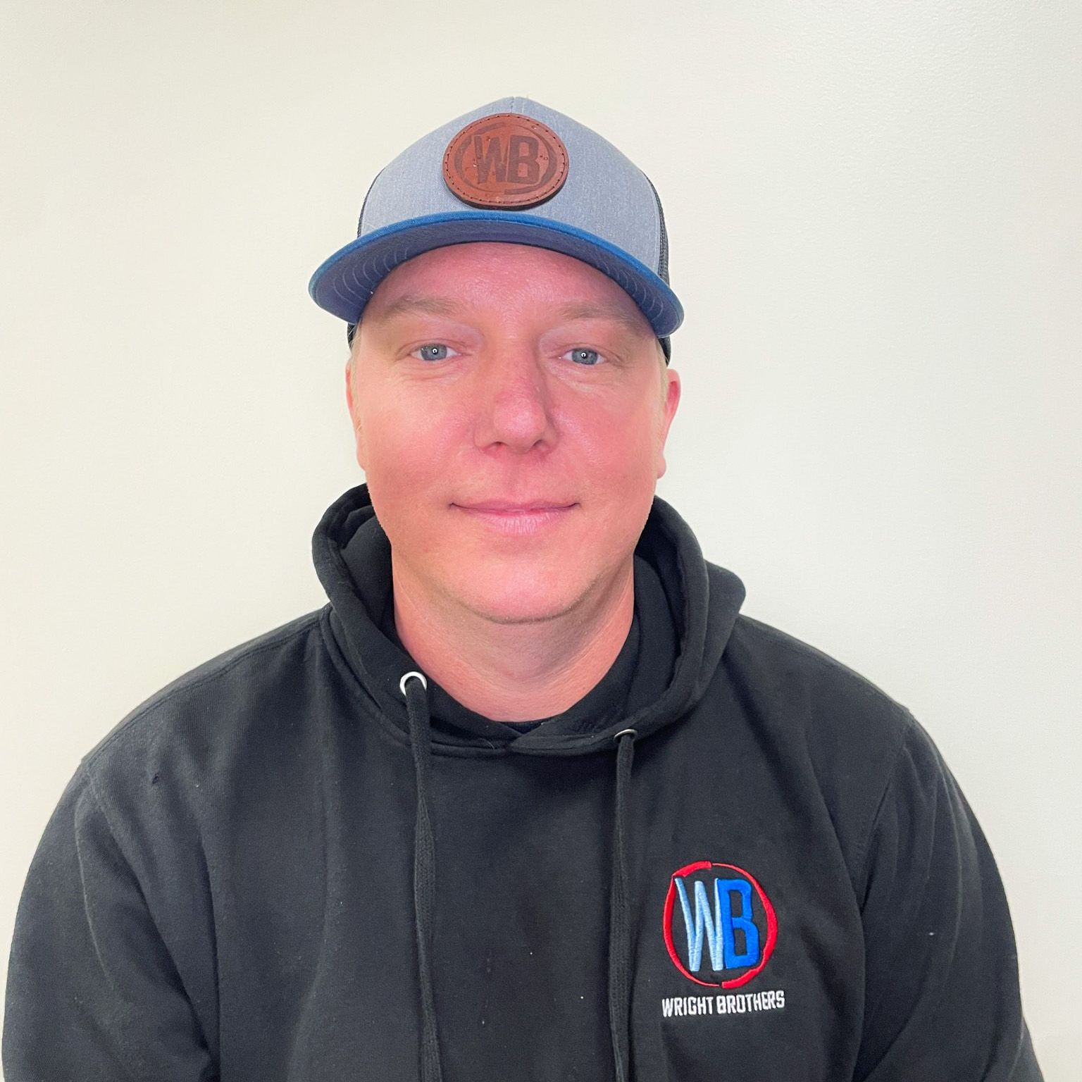 A man wearing a hat and a black hoodie is standing in front of a white wall.