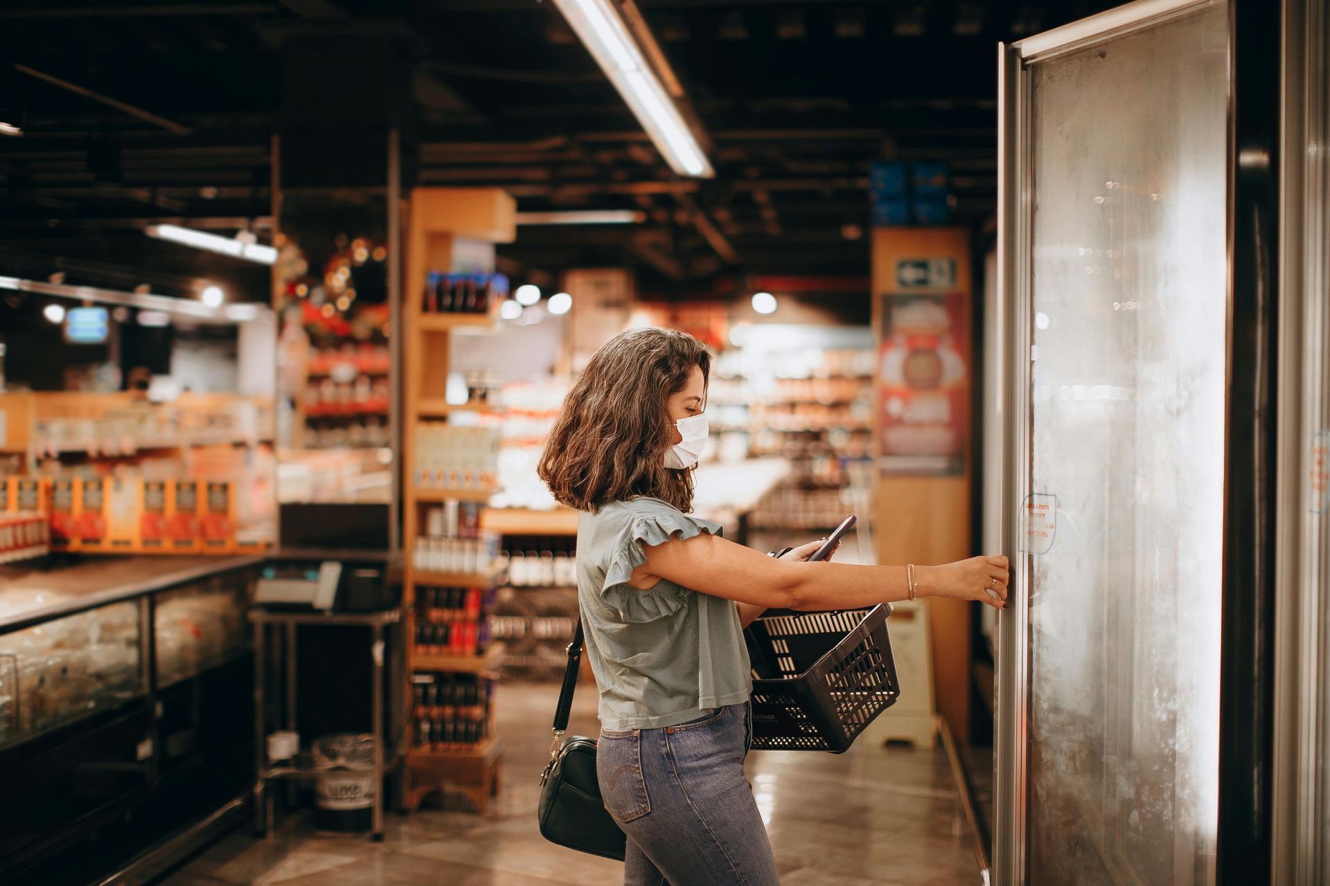 A woman wearing a mask is opening a refrigerator in a supermarket.