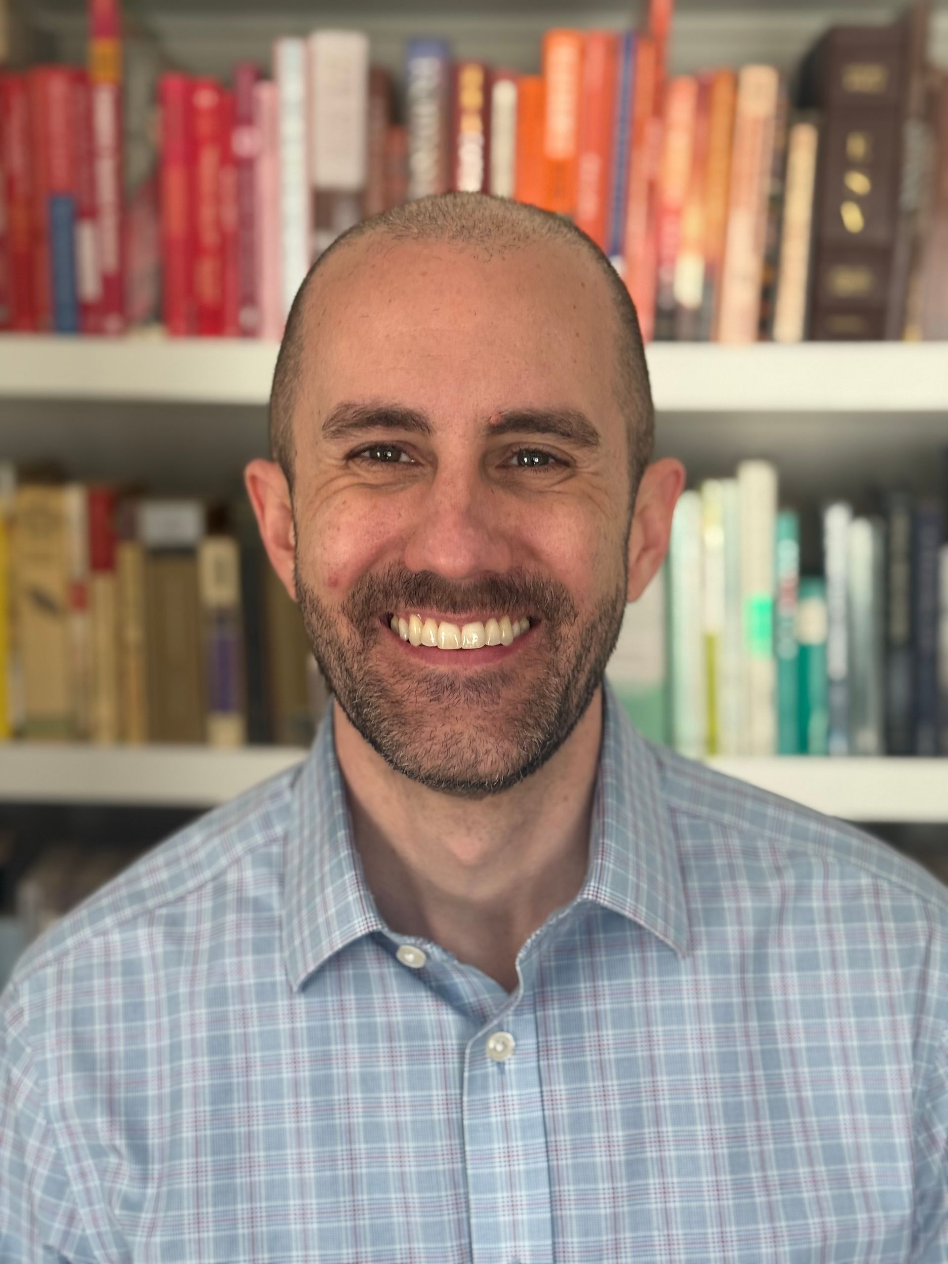 A person with a beard and a light blue checkered shirt smiling in front of a bookshelf.