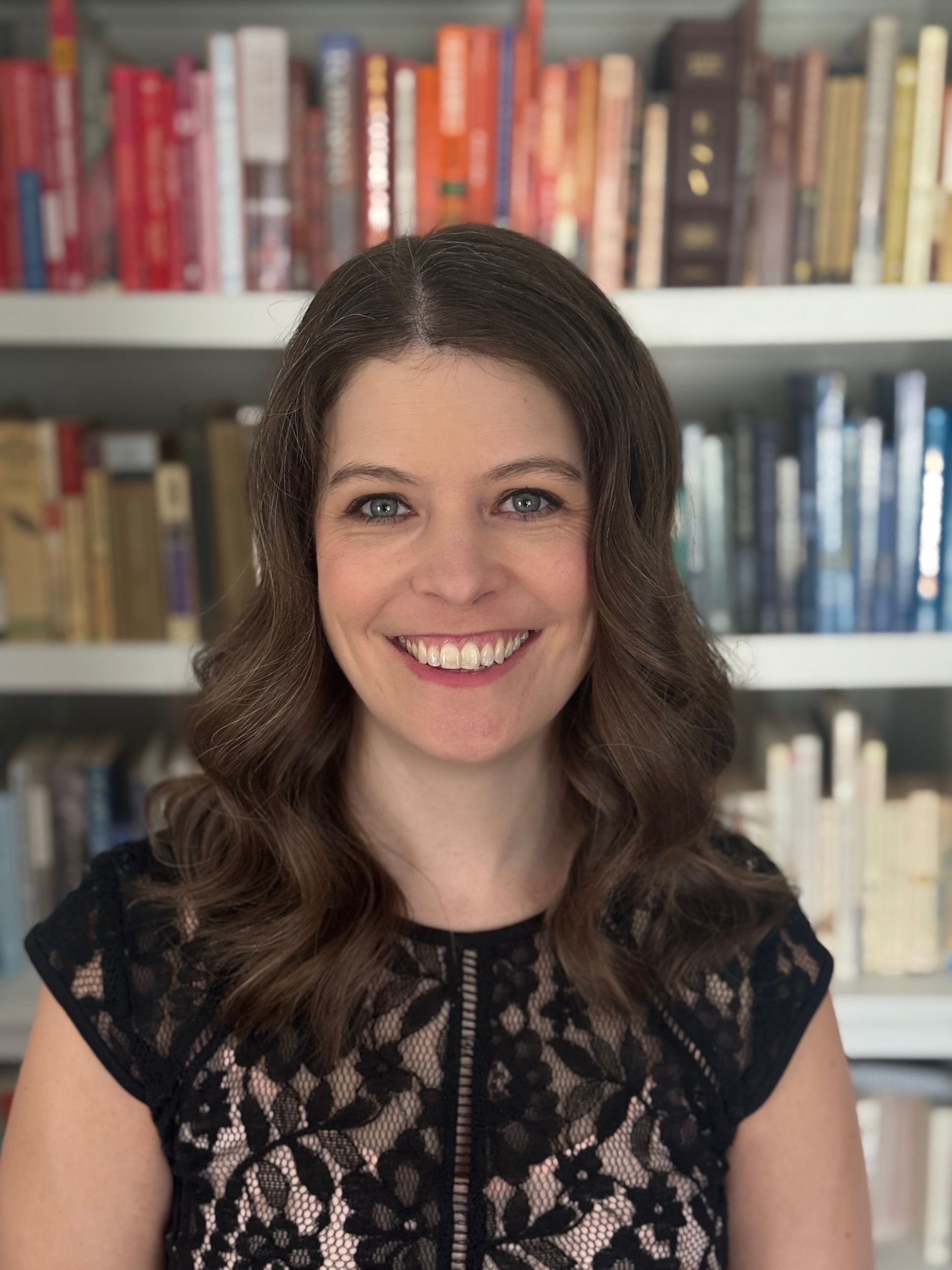 Smiling person with shoulder-length wavy brown hair, wearing a black lace-overlay top in front of a bookshelf.