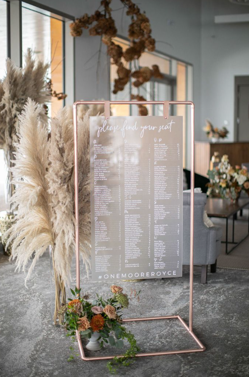 A seating chart is sitting in a room next to pampas grass.