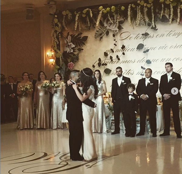 A bride and groom are dancing in front of a wall that says 