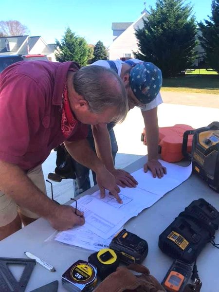 Two workers lean over plans on a white table outdoors, with tools and tape measures nearby.