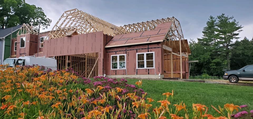 Partially built wooden house with exposed framing beside orange flowers under cloudy sky