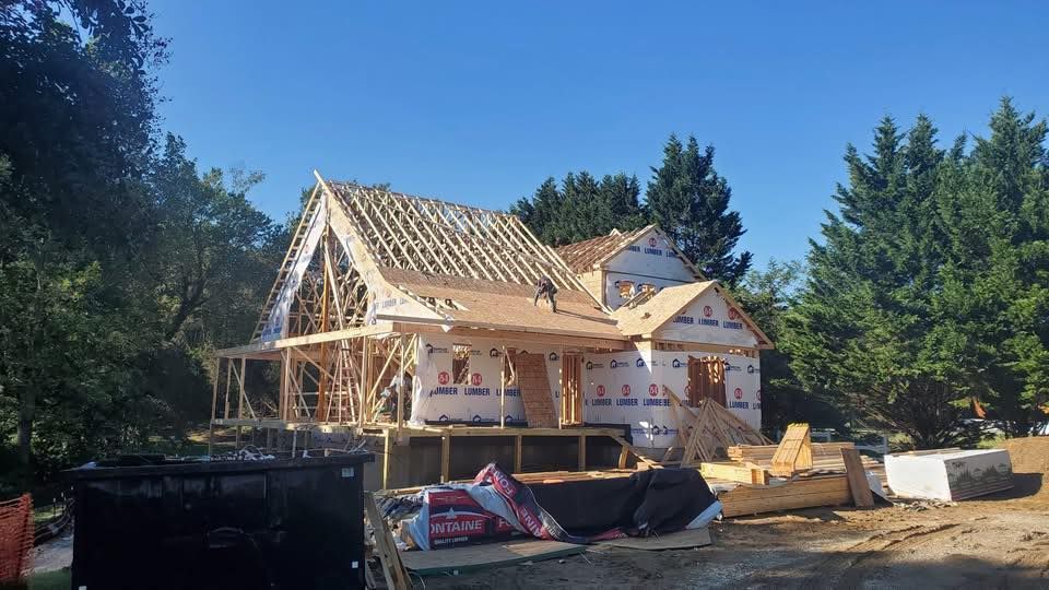 Wood-framed house under construction with exposed rafters, surrounded by trees and building materials.
