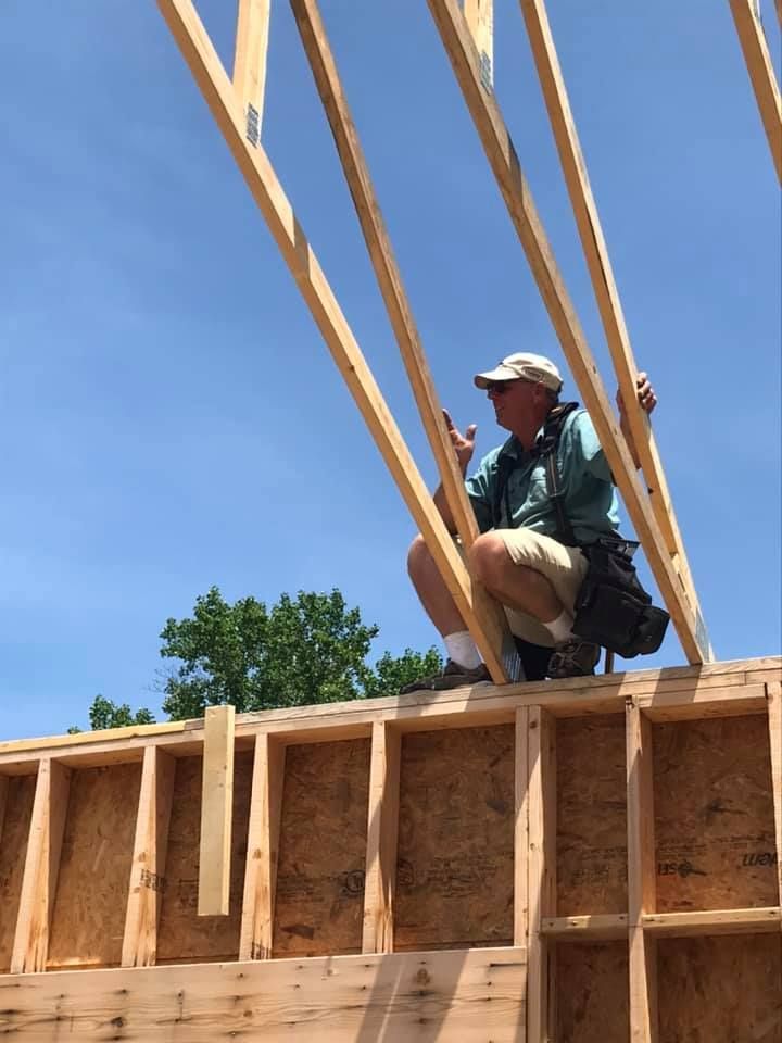 Construction worker kneeling on a framed roof, lifting wooden trusses against a blue sky.