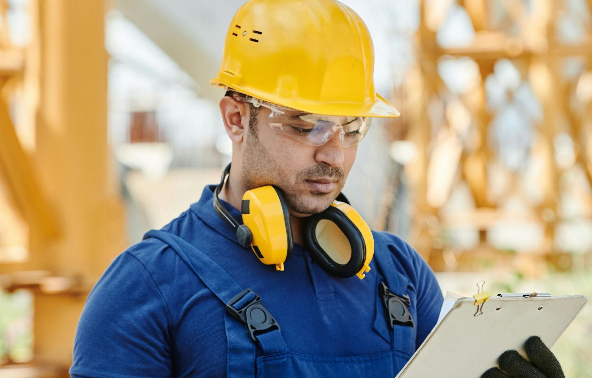 Construction worker in a yellow hard hat and blue coveralls reviewing a clipboard at a work site