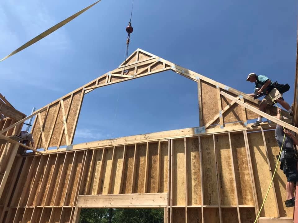Construction workers lifting a wooden roof truss onto a framed building against a blue sky