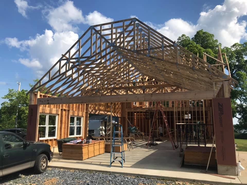 House under construction with exposed wooden roof framing, ladders, and a parked truck outside