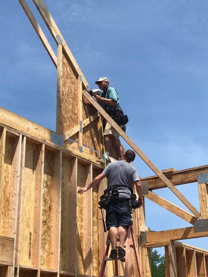 Two construction workers on a wooden frame, one climbing a ladder and one working near a wall under a blue sky