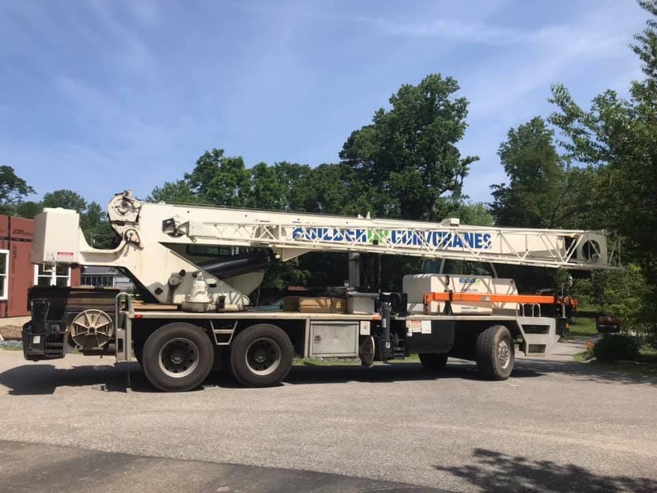 White crane truck parked on a road, with blue lettering and trees in the background.